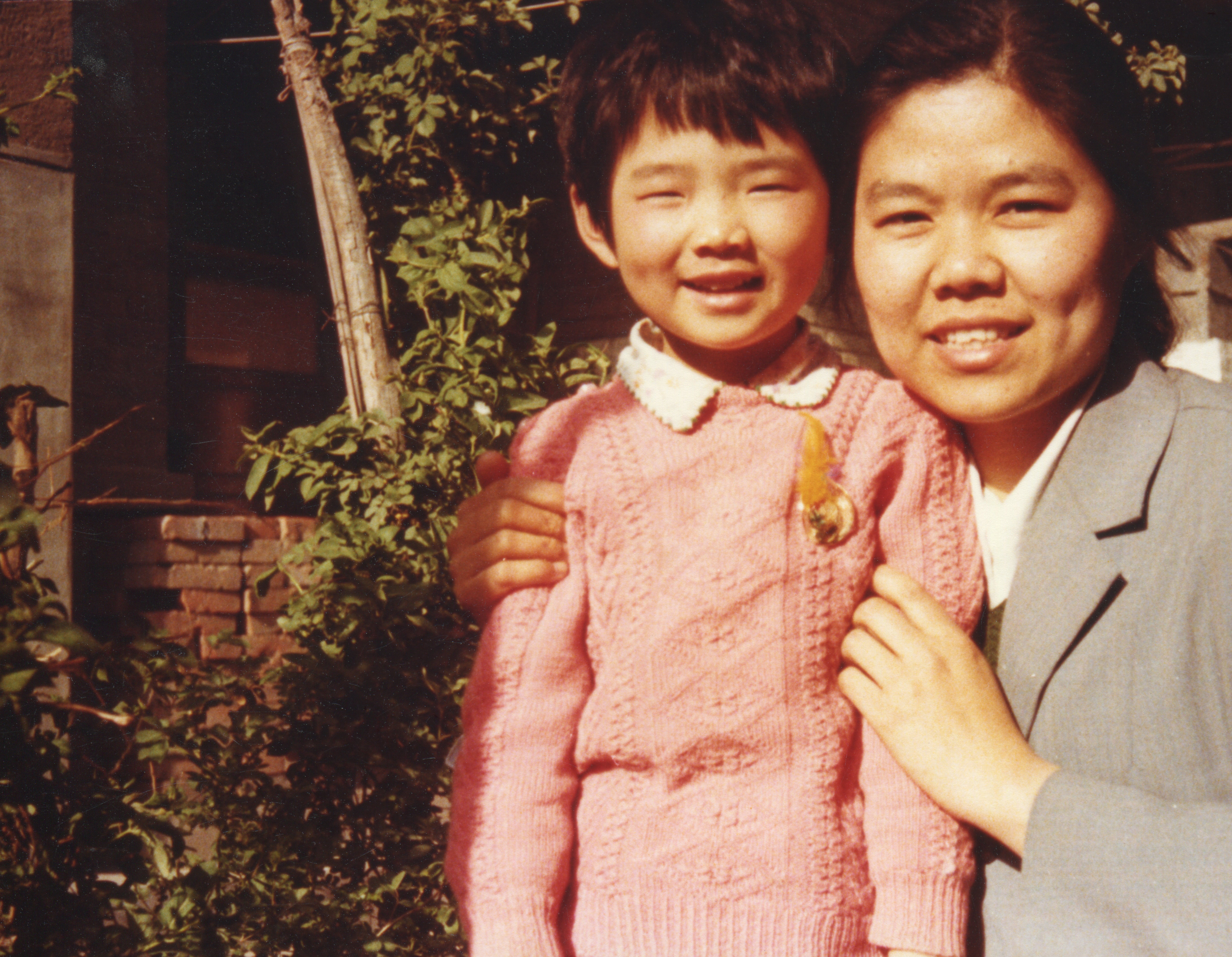 A pistillate   with a gentle grin  holds a smiling kid  wearing a knit sweater with a badge, outdoors by greenery
