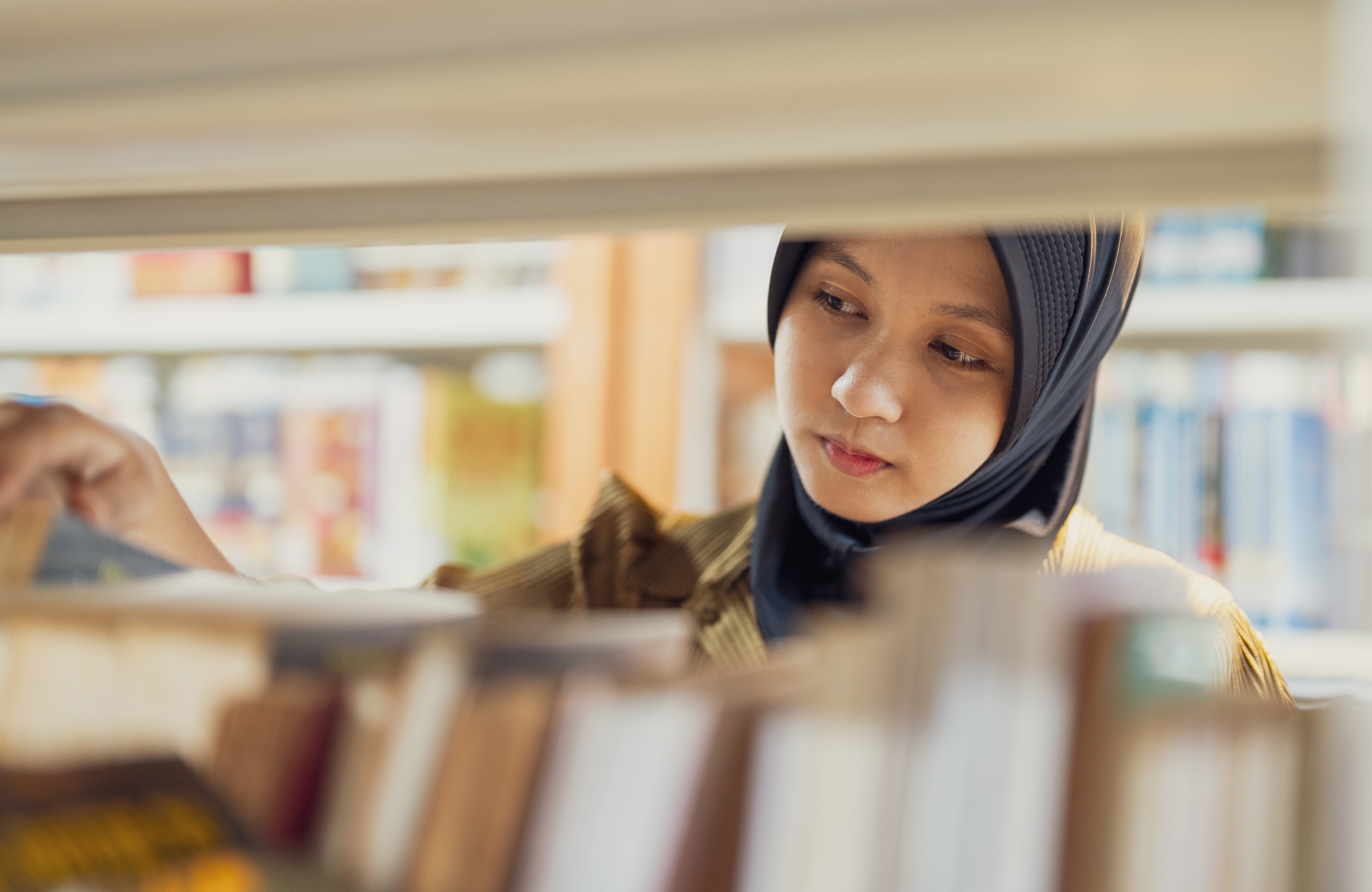 Person successful  a room  browsing books connected  a shelf, wearing a headscarf and looking focused connected  selecting a book