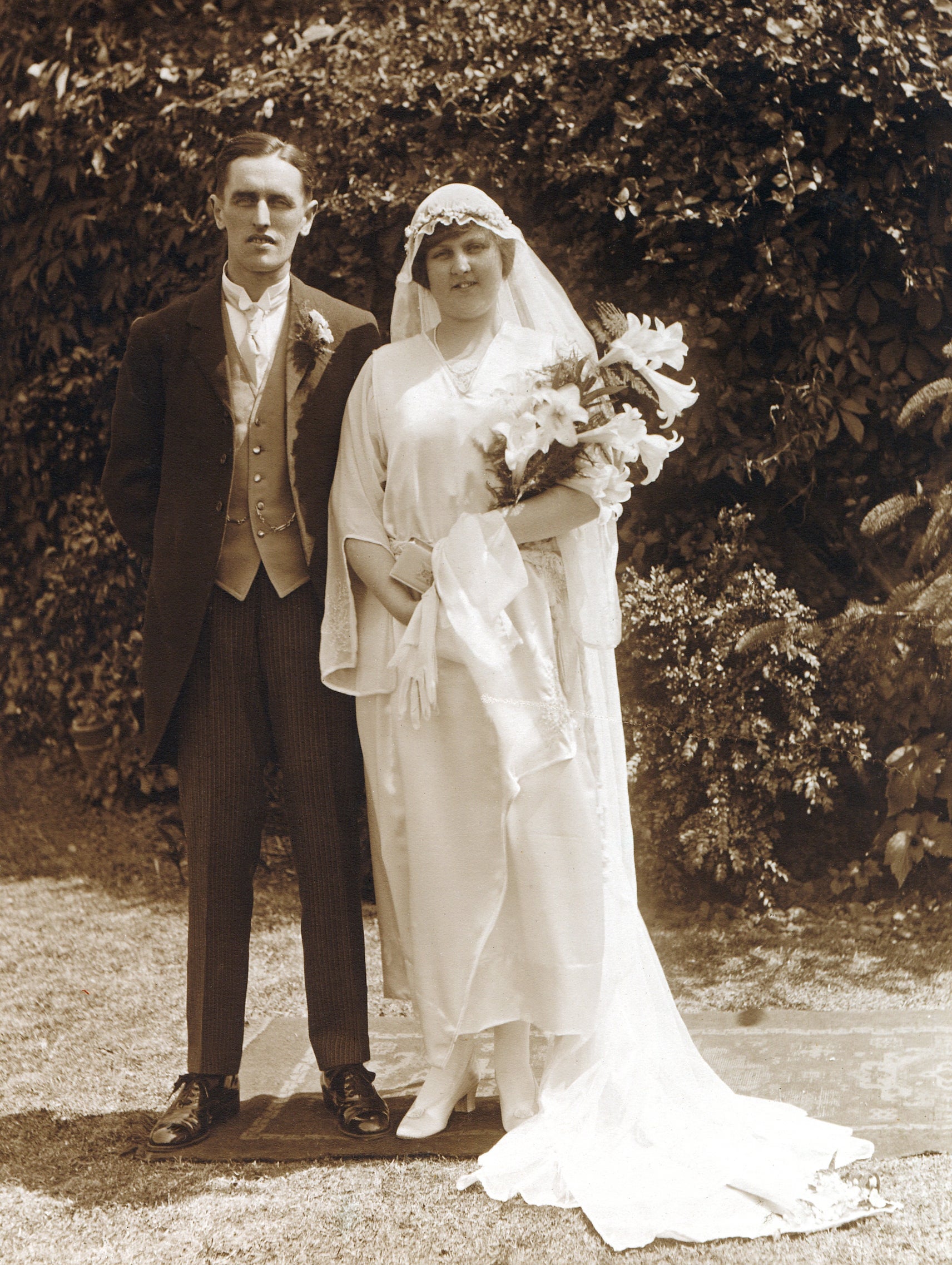 A vintage wedding photograph  of a couple. The groom wears a ceremonial  suit, and the bride is successful  a classical  gown with veil, holding a bouquet of flowers