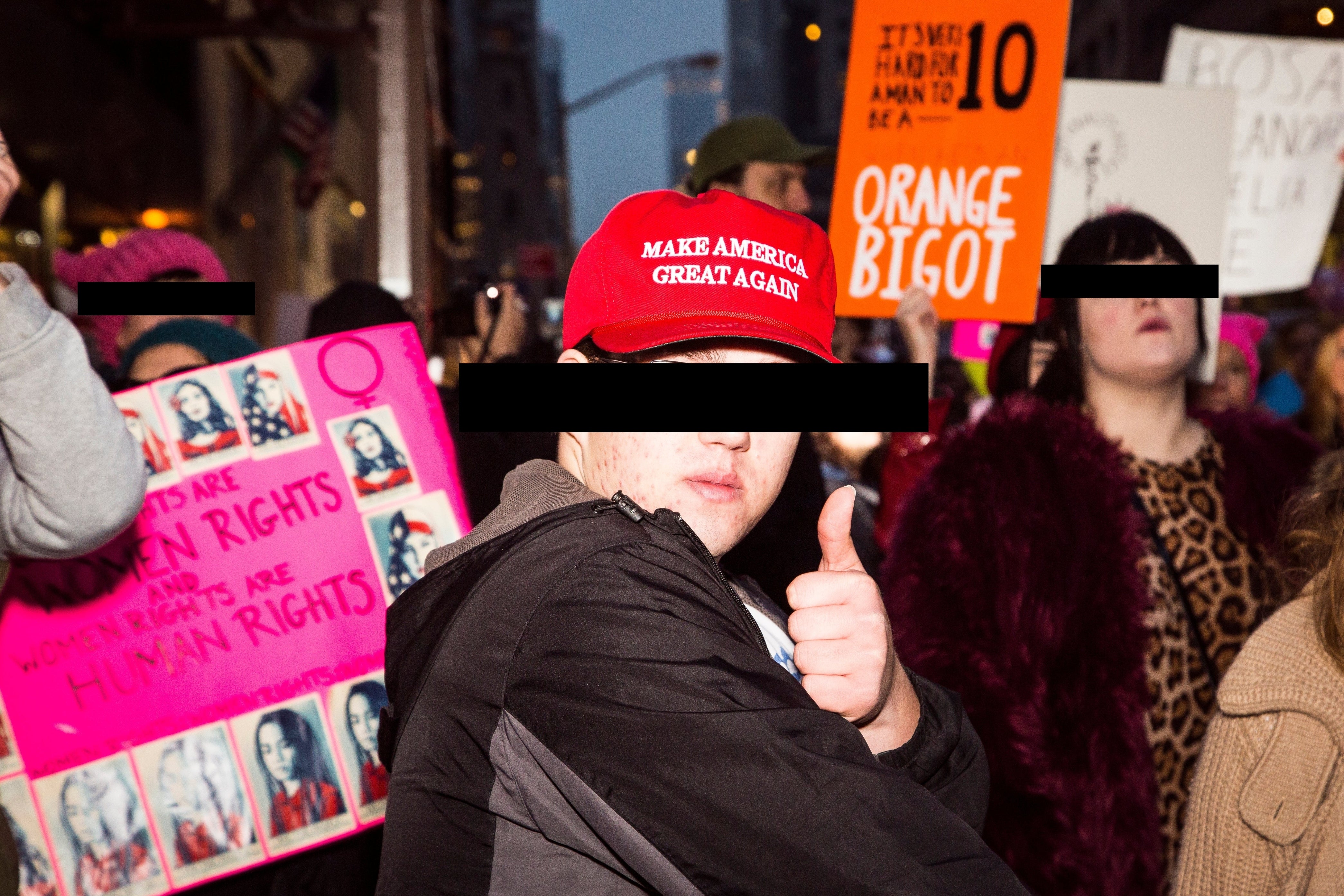 En person som har på seg en "Make America Great Again" cap gir tommel opp midt i en protest med forskjellige skilt synlige i bakgrunnen