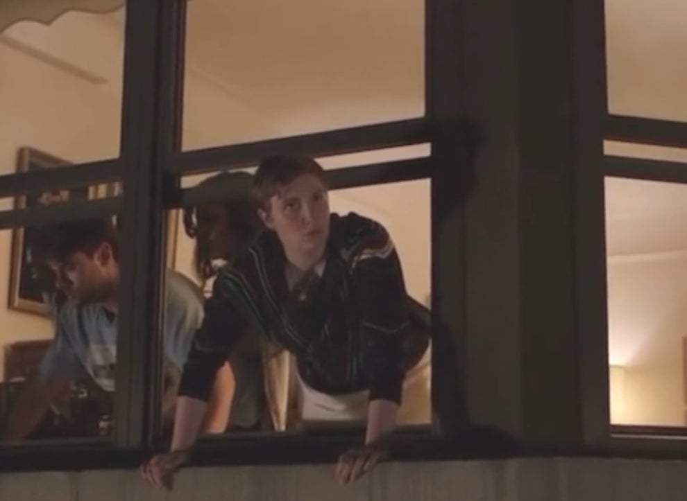 Person leaning out of a window with a thoughtful expression, another person is visible inside the room, partially blurred