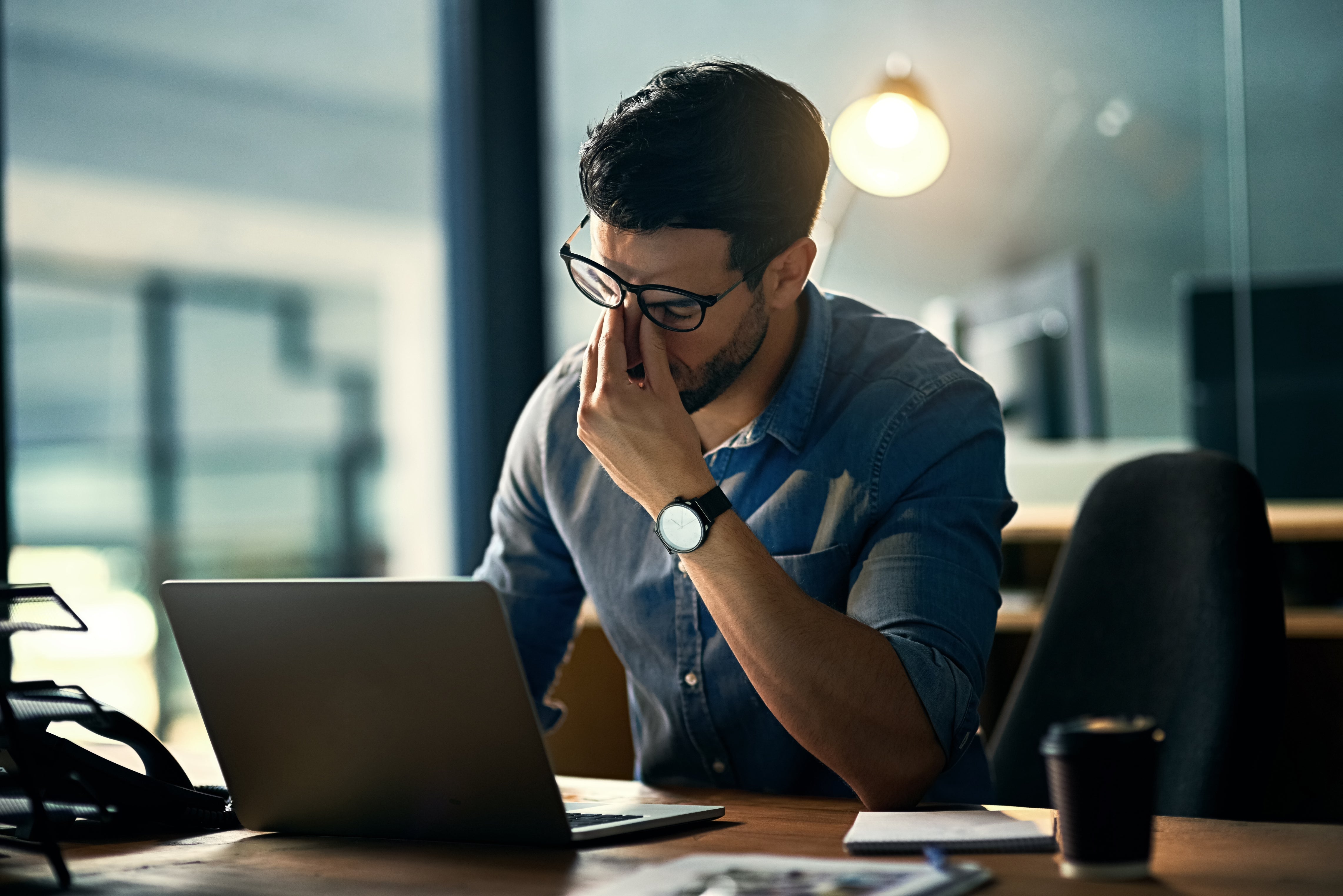 Person successful glasses and casual garment looks tired, sitting astatine a table with a laptop and papers, pinching the span of their nose