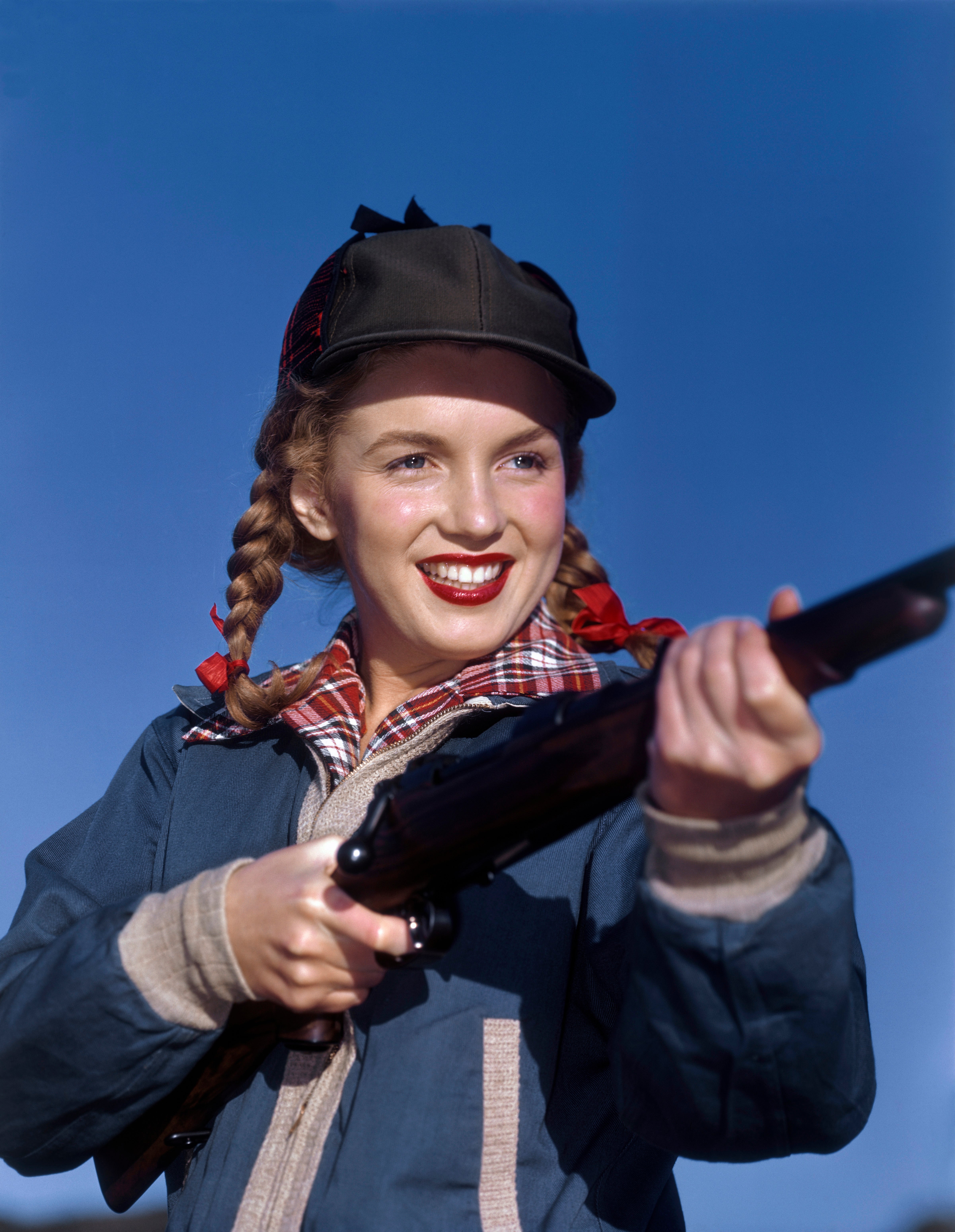 Young Marilyn Monroe with braided hair in a cap, smiling while holding a rifle, outdoors against a clear sky