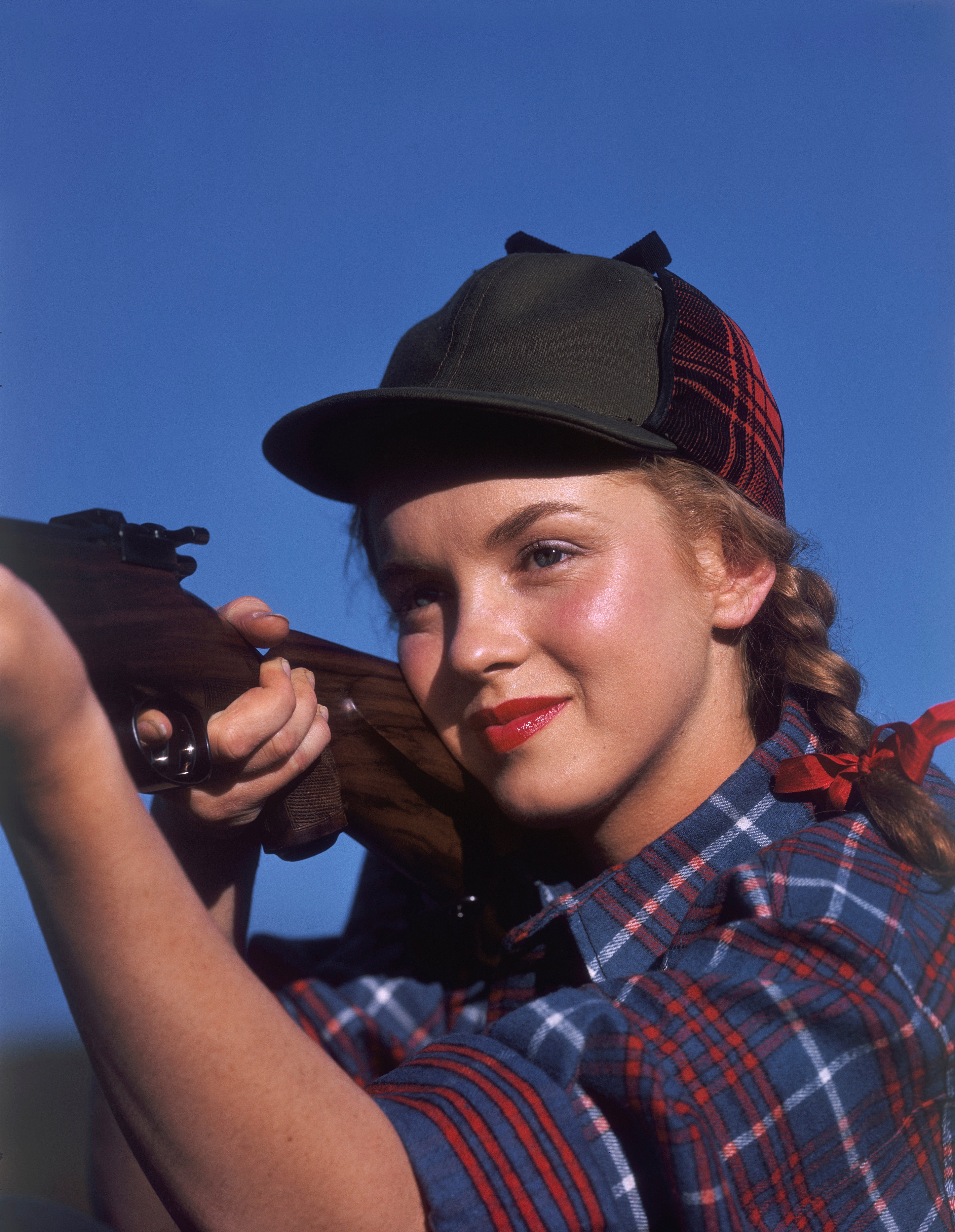 Young Marilyn Monroe with braided hair in a cap, smiling while holding a rifle, outdoors against a clear sky, looking focused