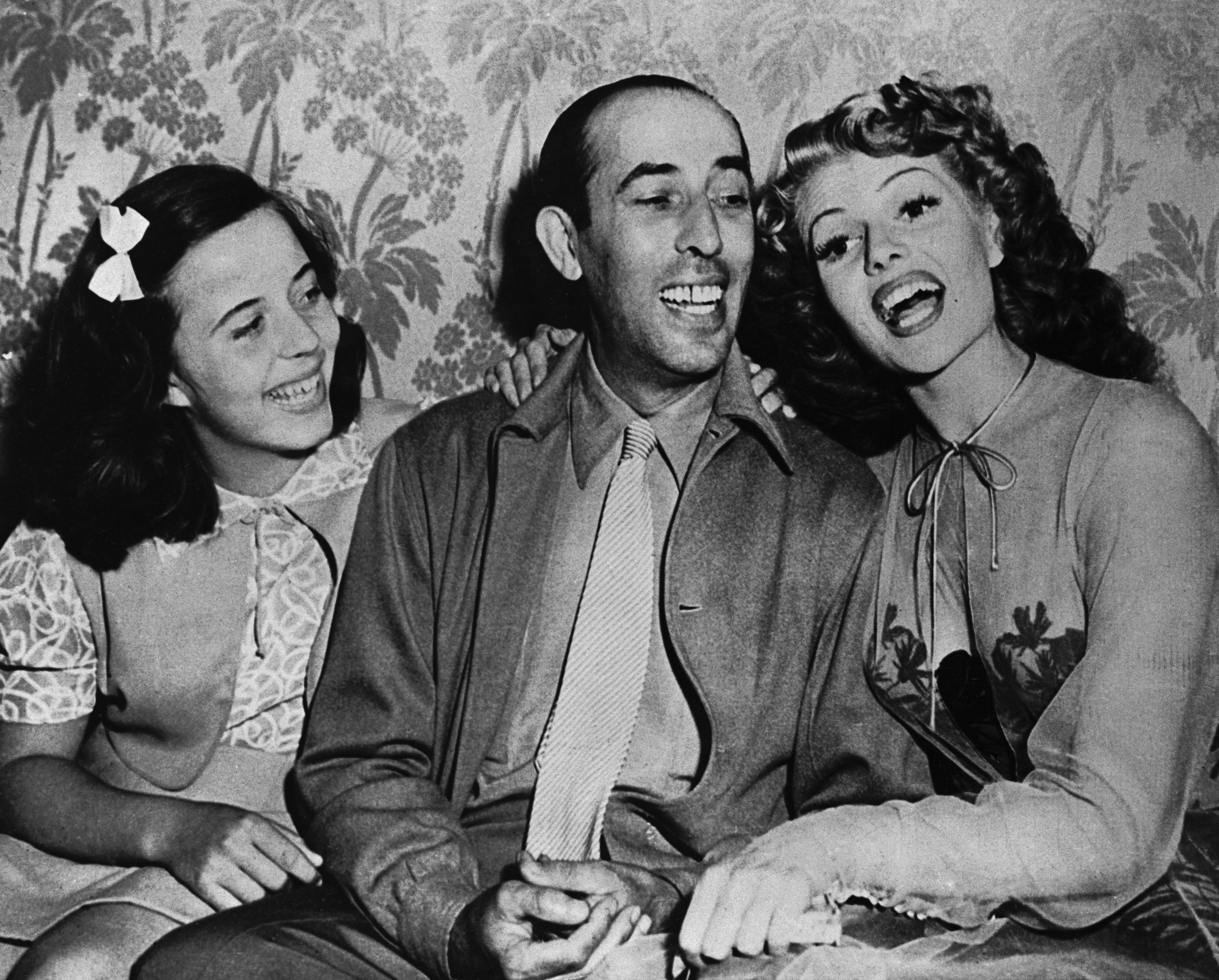 Three people smiling and sitting together, with the two women wearing dresses and the man in a suit and tie. The woman on the right is Rita Hayworth.