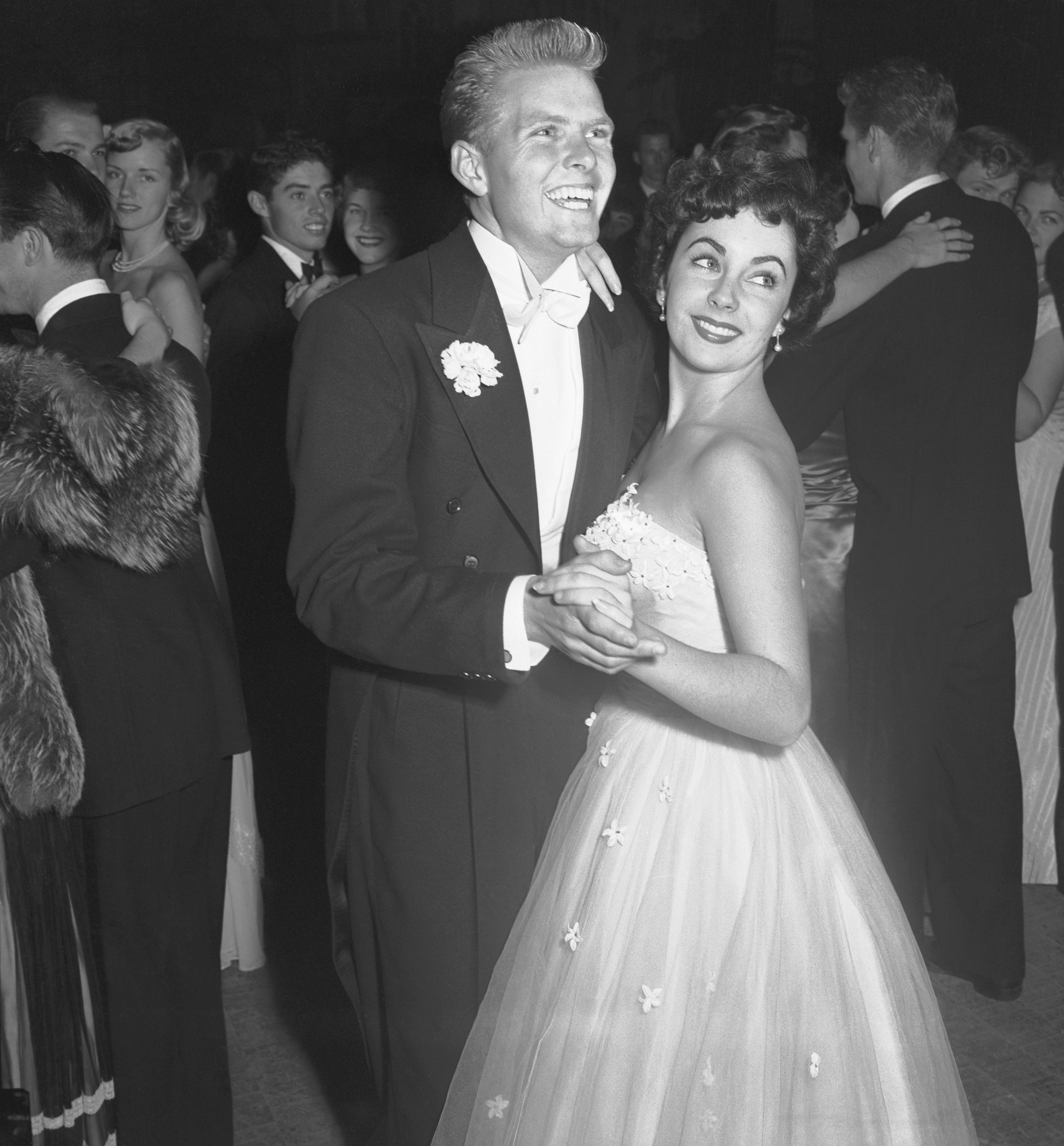 Bob Precht and Elizabeth Taylor in elegant formal wear dancing at a vintage event, surrounded by others in evening attire.