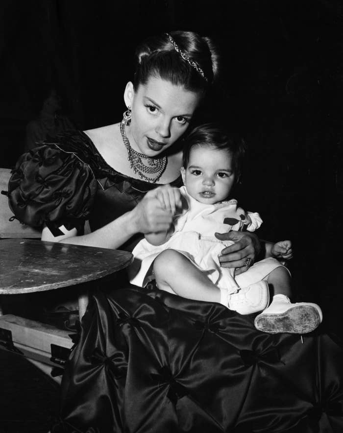 Judy Garland in an elegant gown with a formal hairstyle holds baby Liza Minelli on her lap. She wears a necklace and earrings