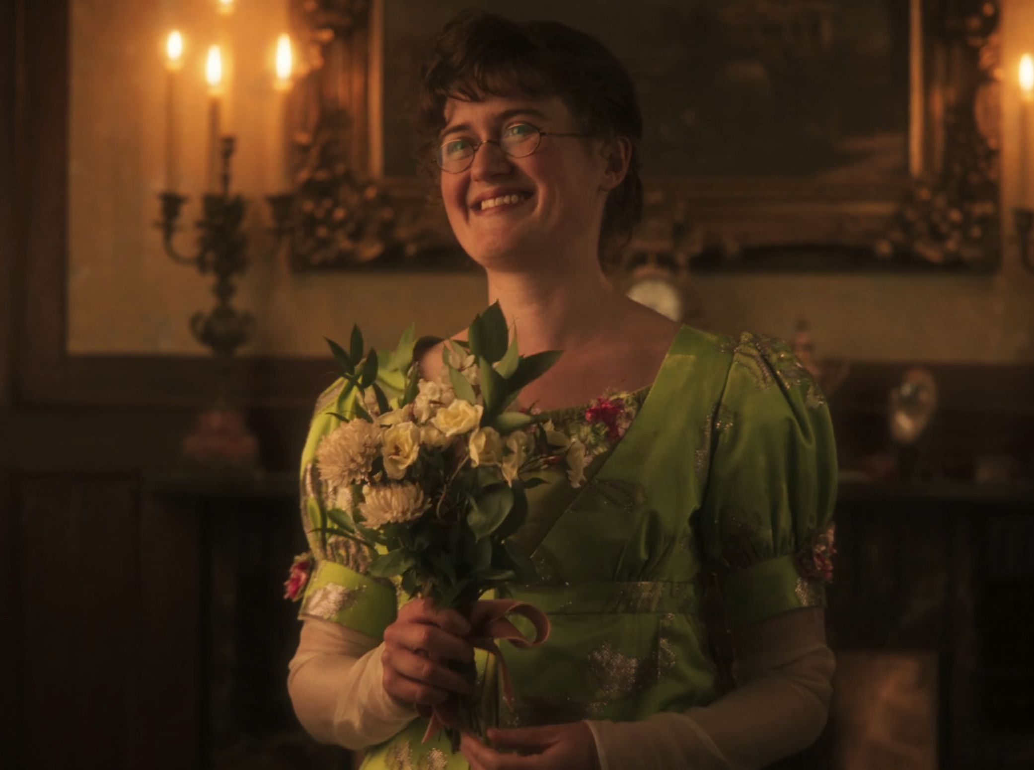 Person in a vintage-style gown, holding a bouquet, smiling warmly in an elegant room with candlelit ambiance