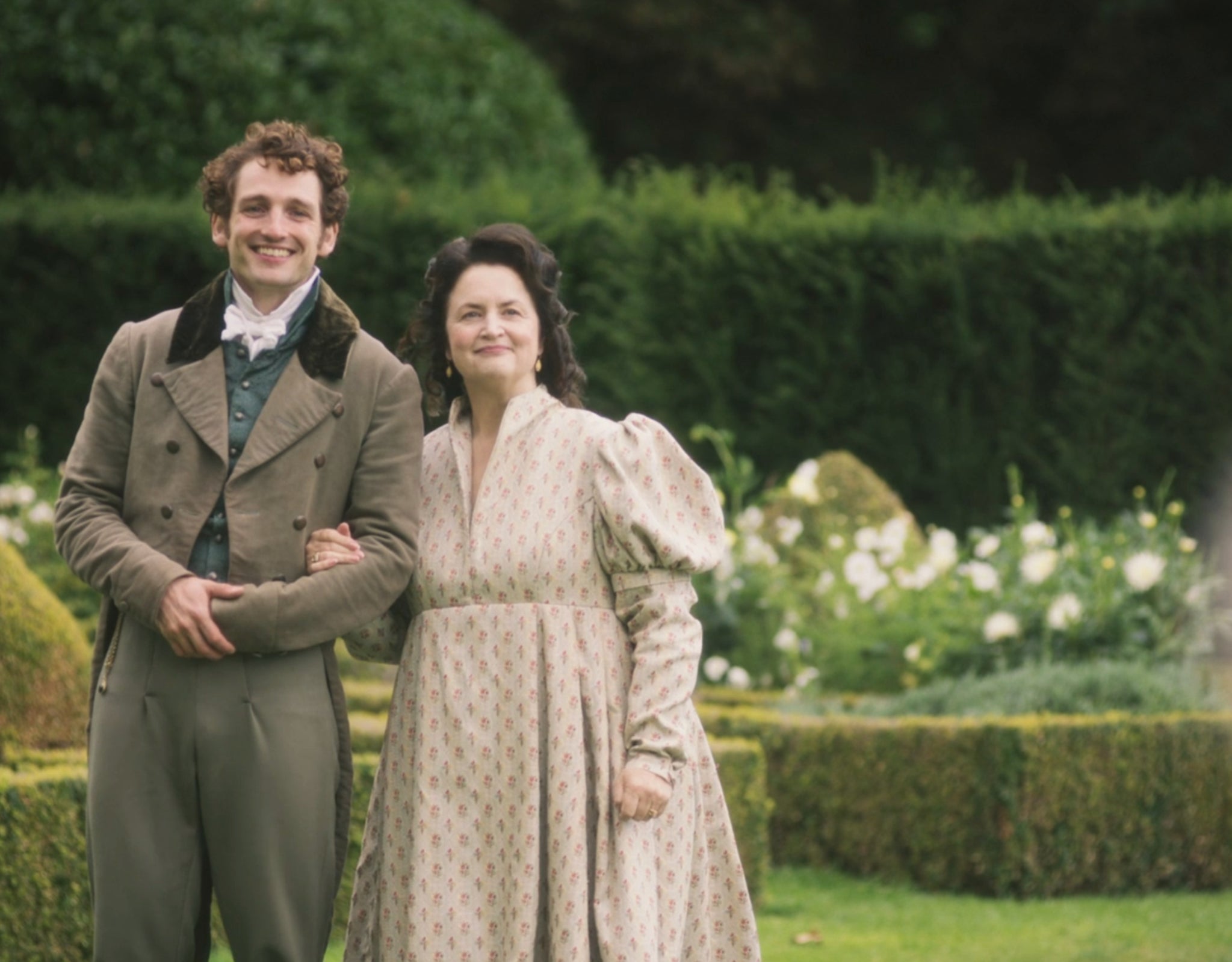 A couple in period clothing stands facing each other in a formal garden, reminiscent of a historical drama setting