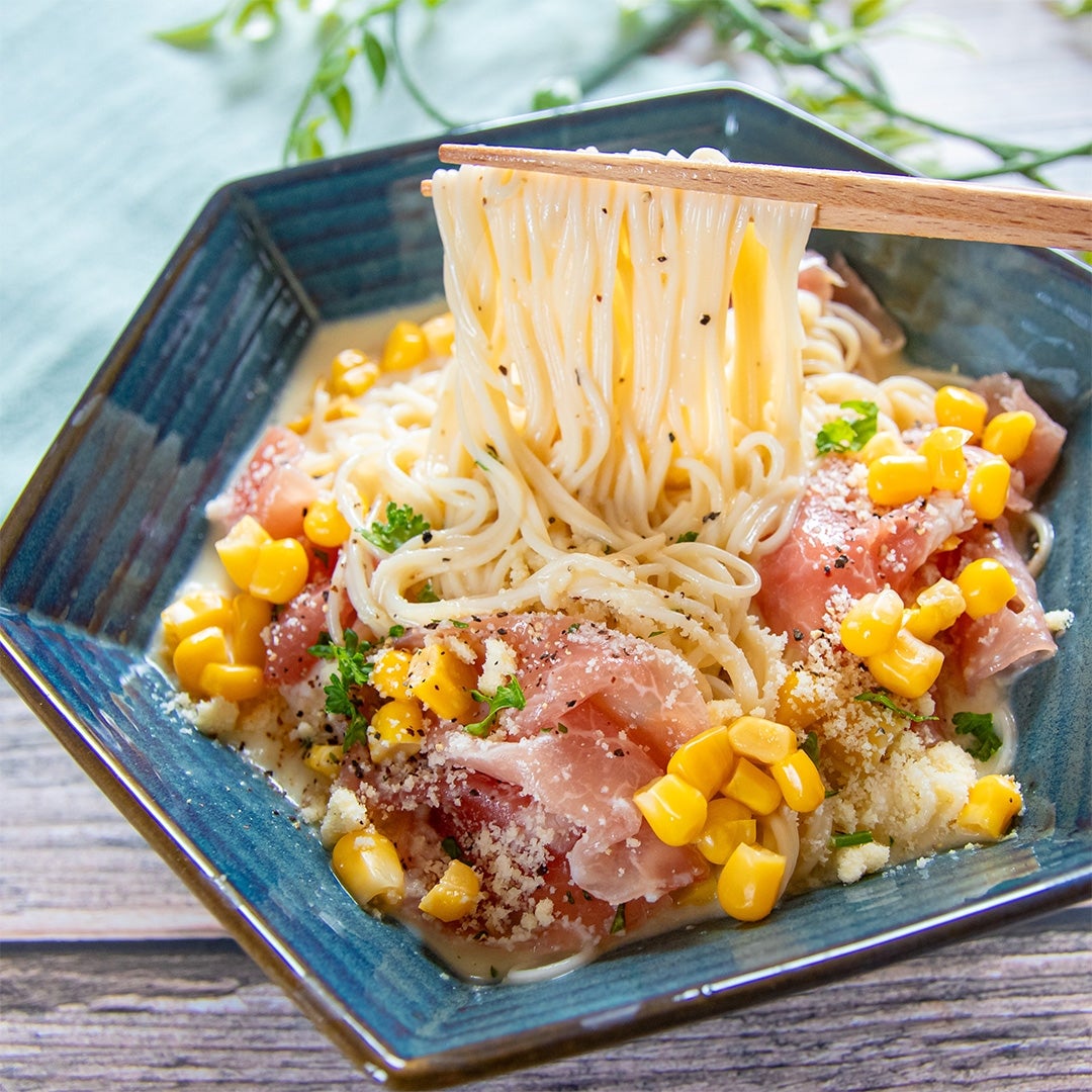 器に盛られたそうめん。コーン、生ハム、パルメザンチーズがトッピングされ、箸で麺を持ち上げている。