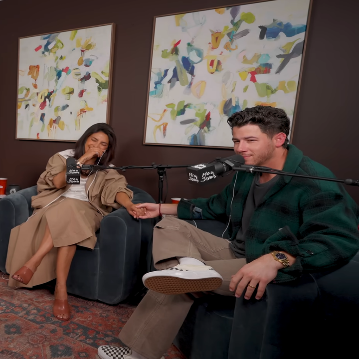 Two people sit in a podcast studio holding hands, microphones in front of them, with abstract paintings in the background