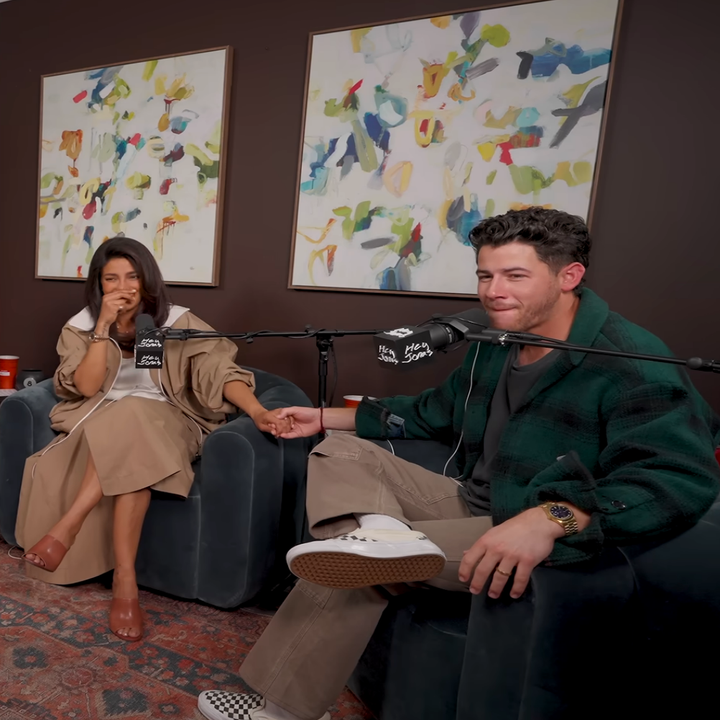 Two people sit on a sofa during a podcast recording. One wears a knee-length coat and the other a plaid jacket. Abstract art is in the background