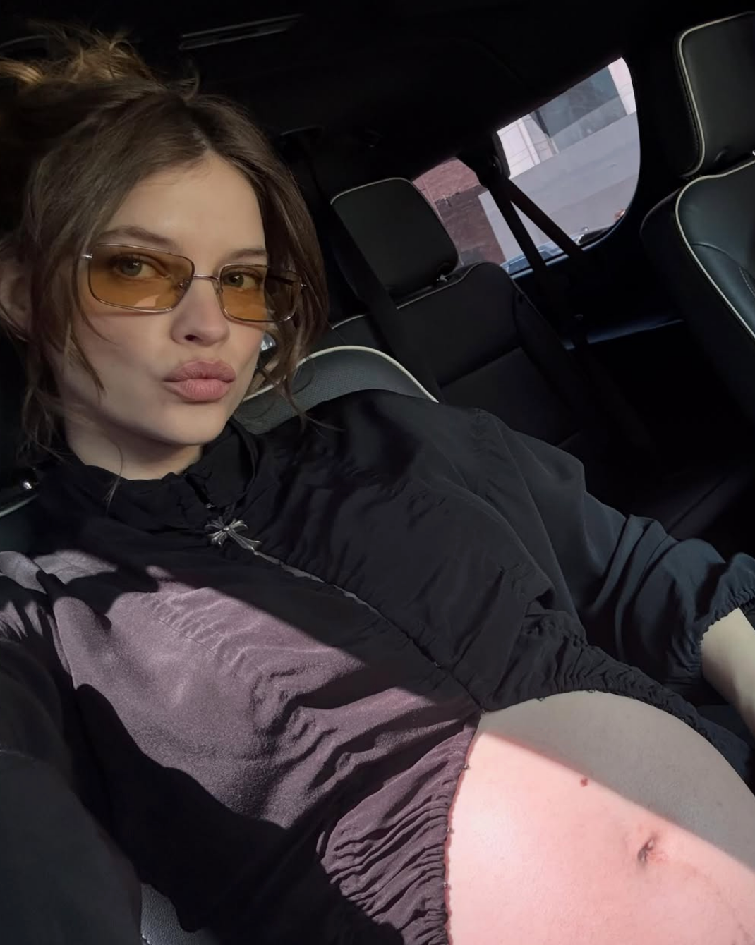 Person posing in a car, visible baby bump, wearing sunglasses and a casual top, conveying relaxed style