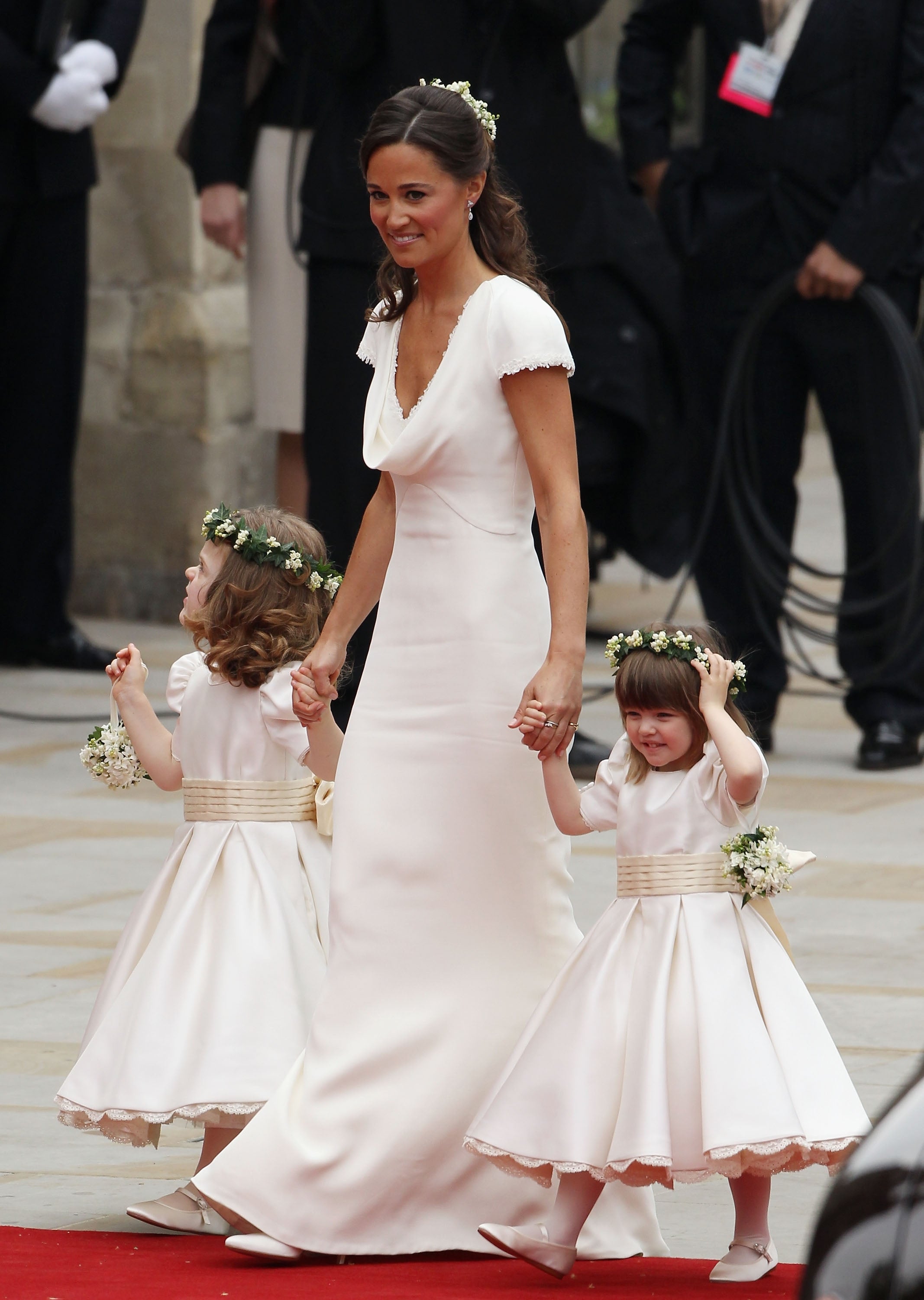A woman in a simple, elegant gown walks with two flower girls in matching dresses and floral headbands at a formal event