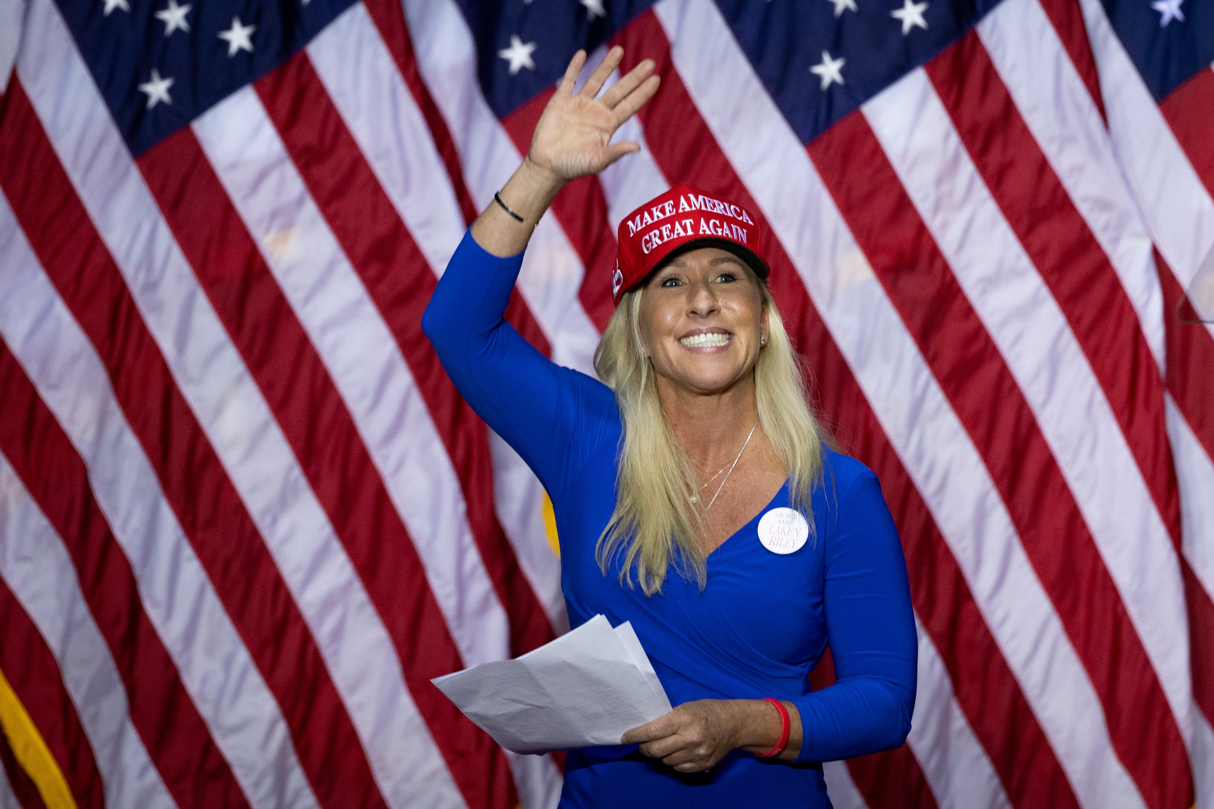 Person i blå kjole og "Make America Great Again" hattebølger, holder papirer, står foran amerikanske flagg