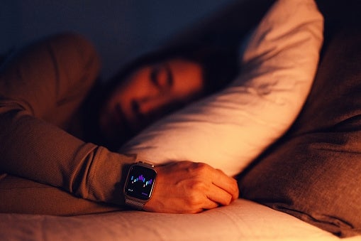 A idiosyncratic sleeps with a smartwatch connected their wrist, displaying a graph. They remainder connected a pillow, suggesting slumber tracking technology