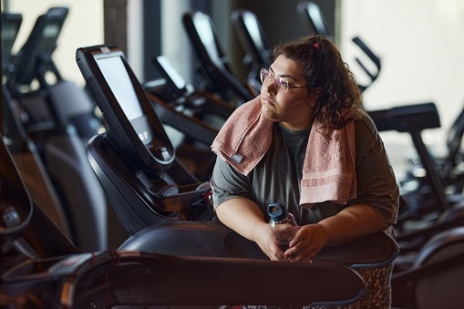 Person with a towel astir their cervix resting connected gym equipment, holding a h2o bottle, and looking thoughtfully into the distance
