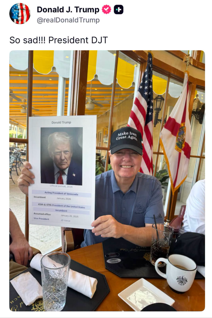 A man at a table holds a framed image of Donald Trump labeled as a fictional acting president, smiling, wearing a "Make America Great Again" cap