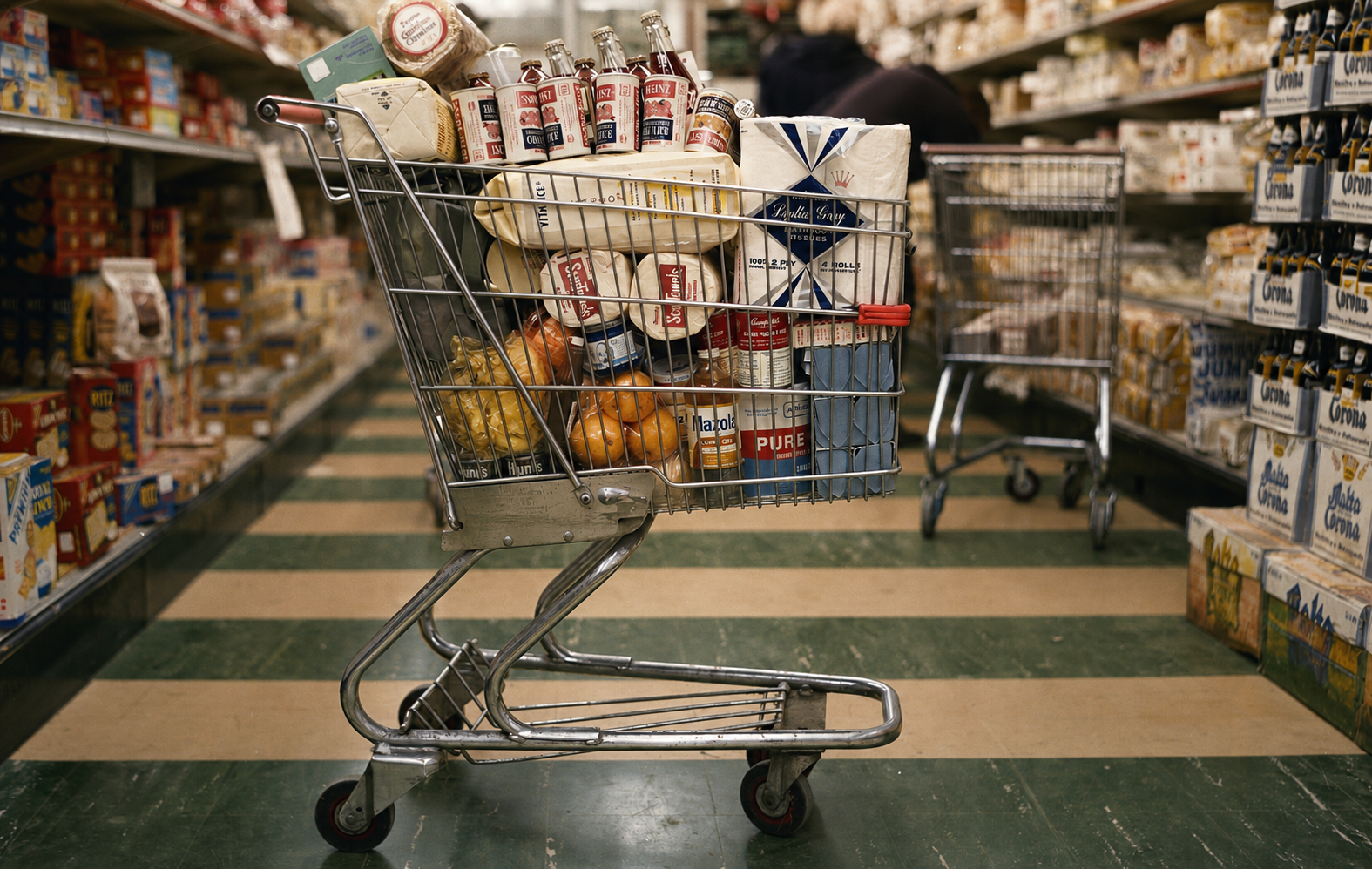 Shopping cart filled with various groceries, including canned goods, vegetables, and packaged items, in a supermarket aisle