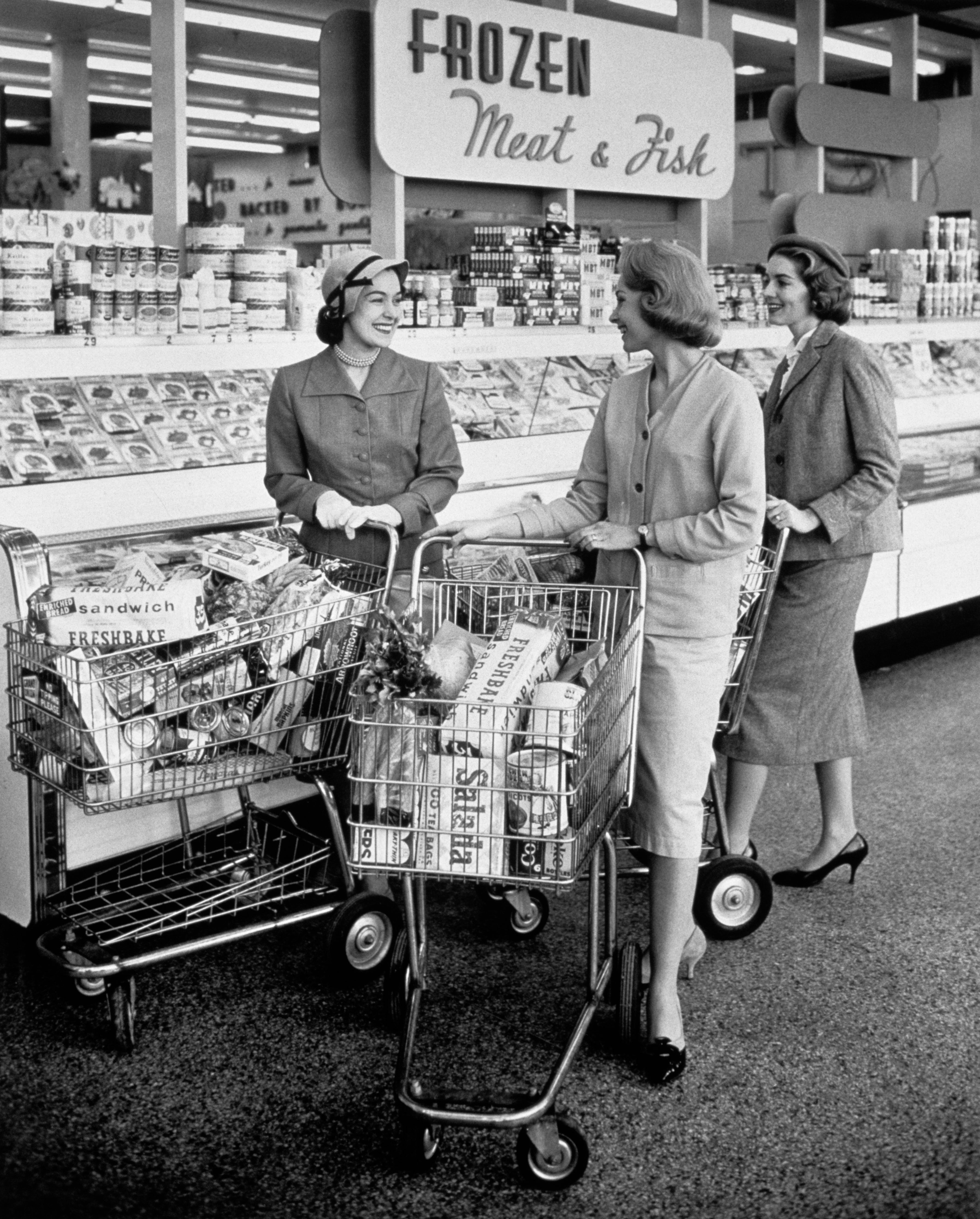 Three women in 1950s attire chat while shopping in a grocery store aisle with carts full of groceries, near a sign for frozen meat and fish