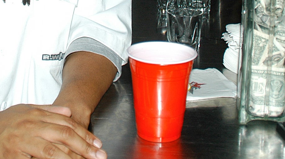 Person with dreadlocks wearing sunglasses and a white shirt with a logo stands at a bar with a red cup and bottles, surrounded by people
