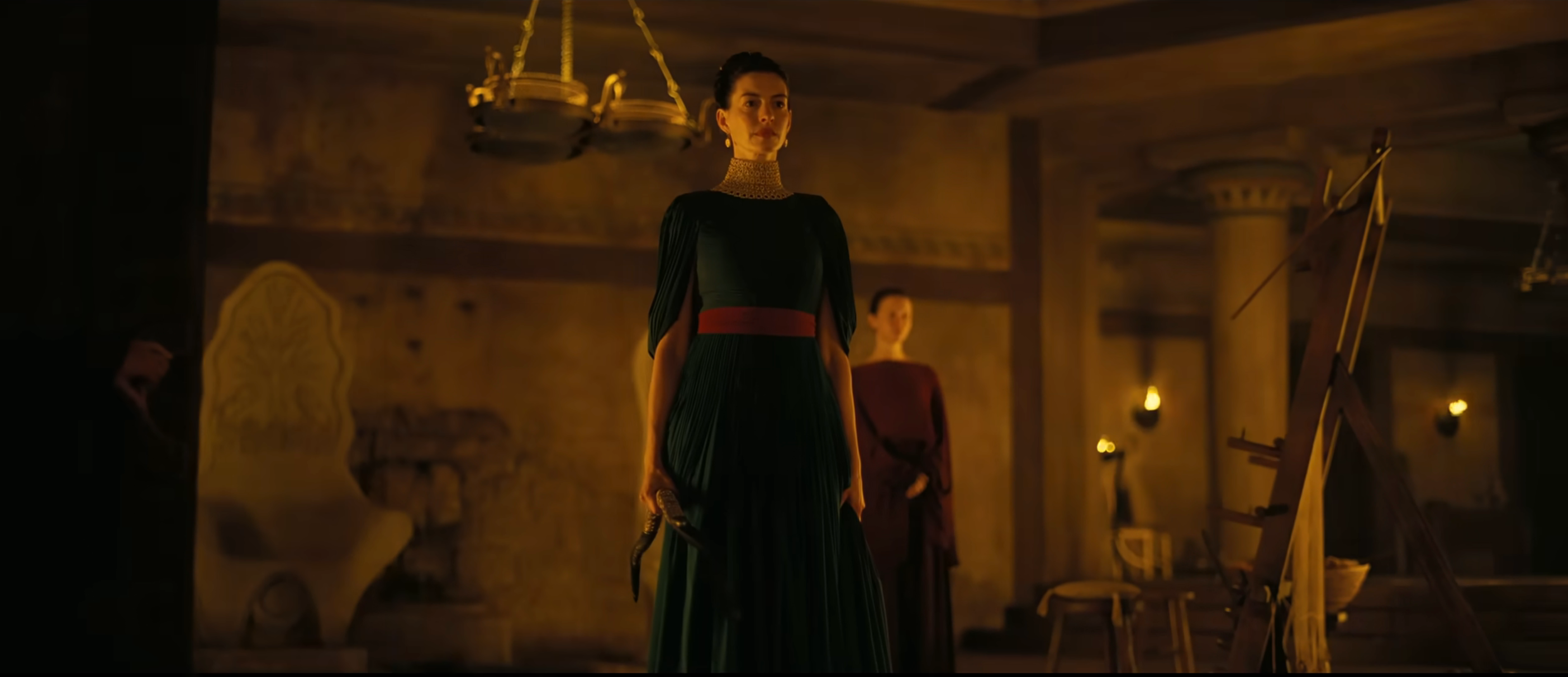 A woman in elegant period clothing stands in a dimly lit room with classical decor, evoking a historical or dramatic scene