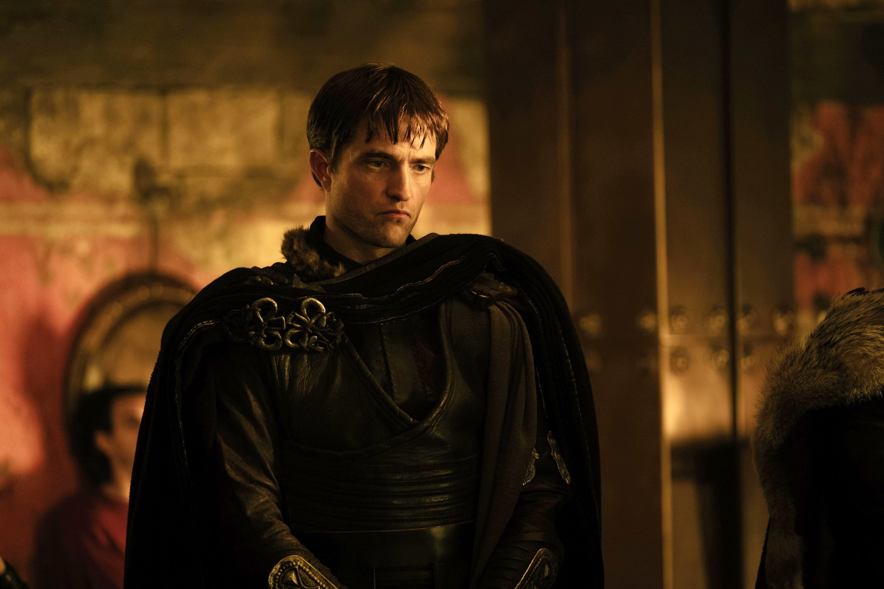 An actor in medieval-style armor and cloak, standing in a dimly lit room