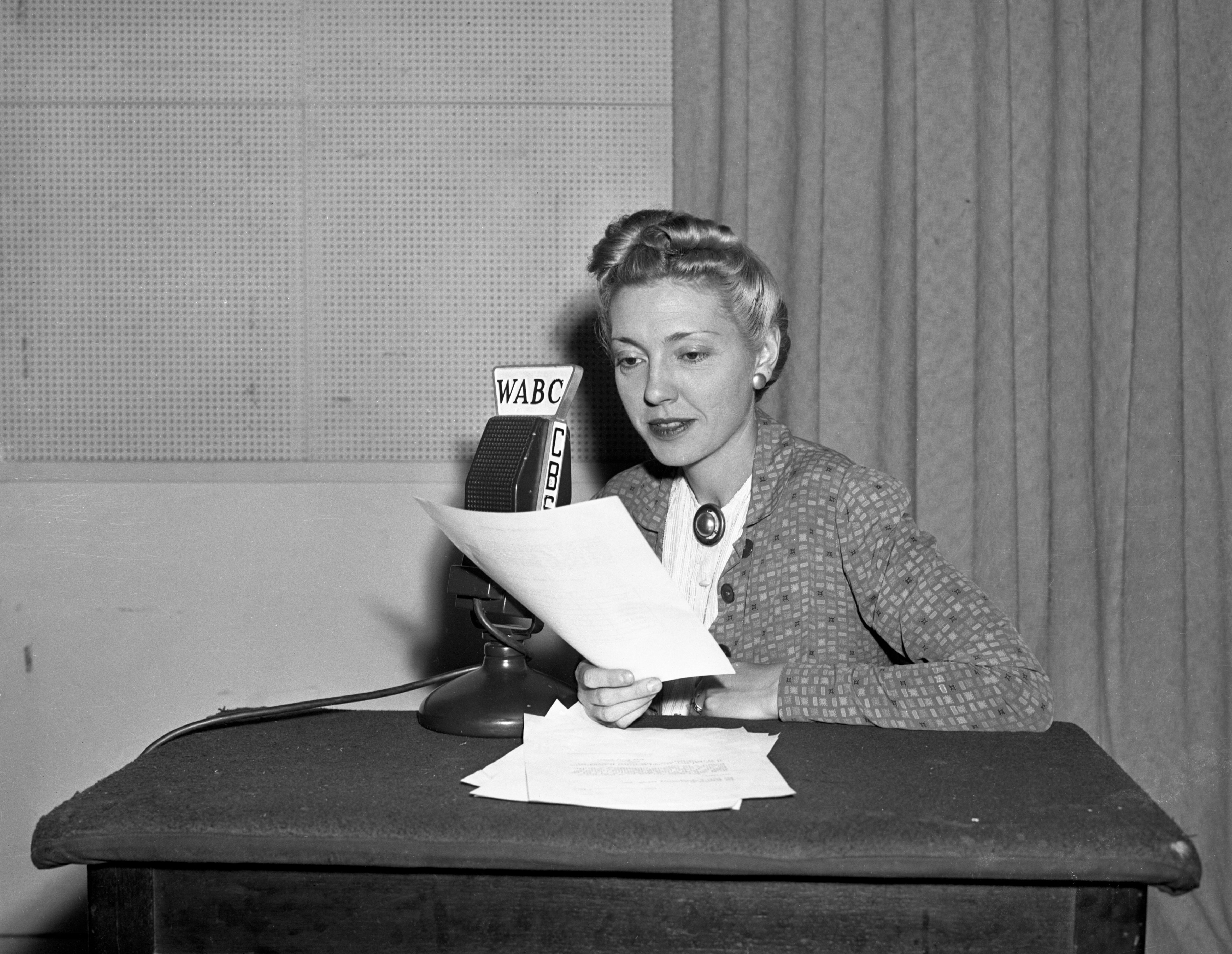 Woman in vintage attire speaking into a WABC radio microphone at a desk with papers, likely from an earlier era in broadcasting