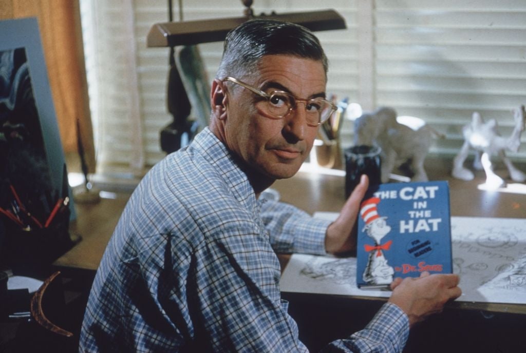 Dr. Seuss seated at a desk holding a copy of "The Cat in the Hat" book, glancing back at the camera