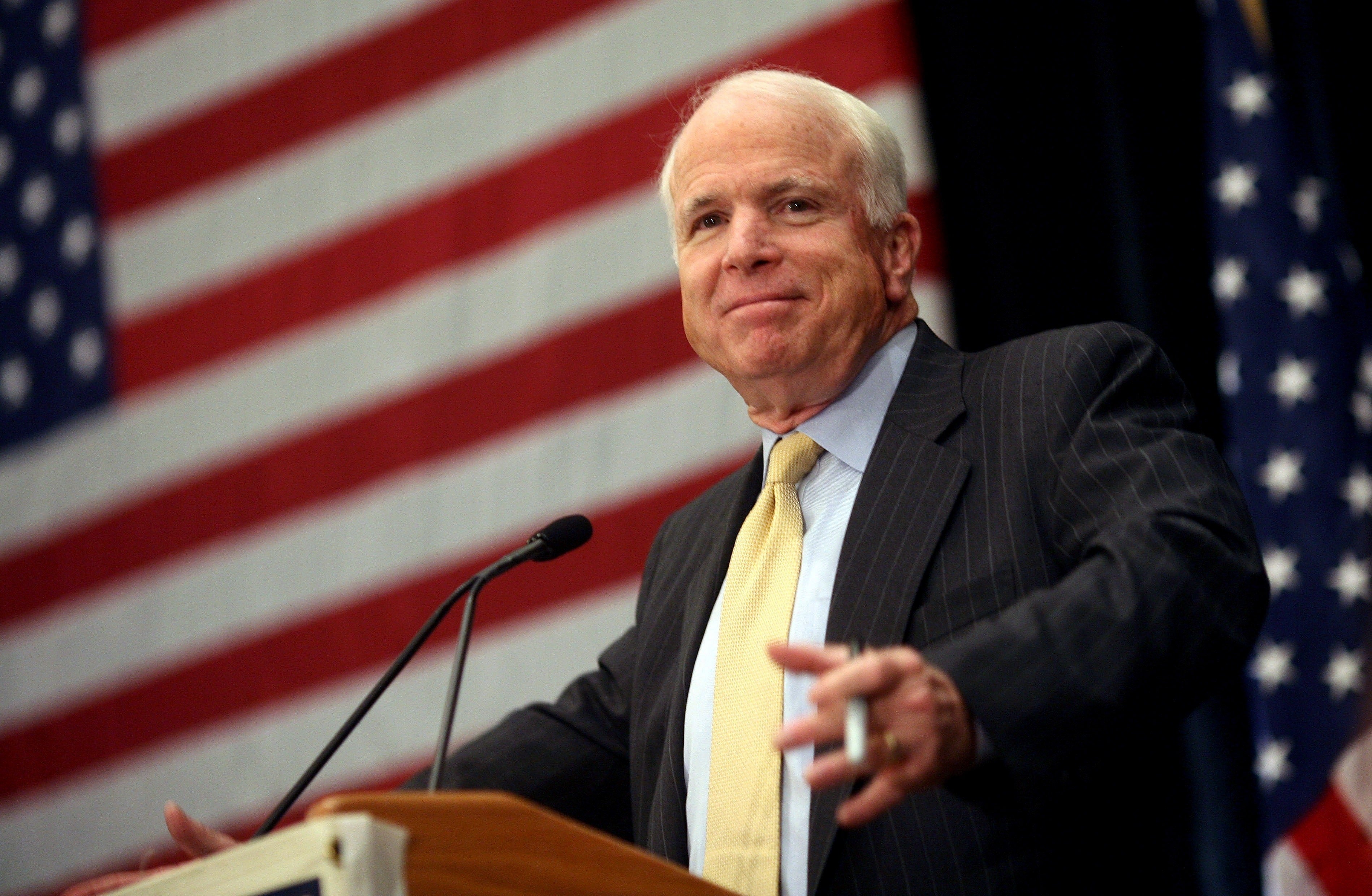 A person in a suit speaks at a podium with U.S. flags in the background, appearing at a public event or press conference
