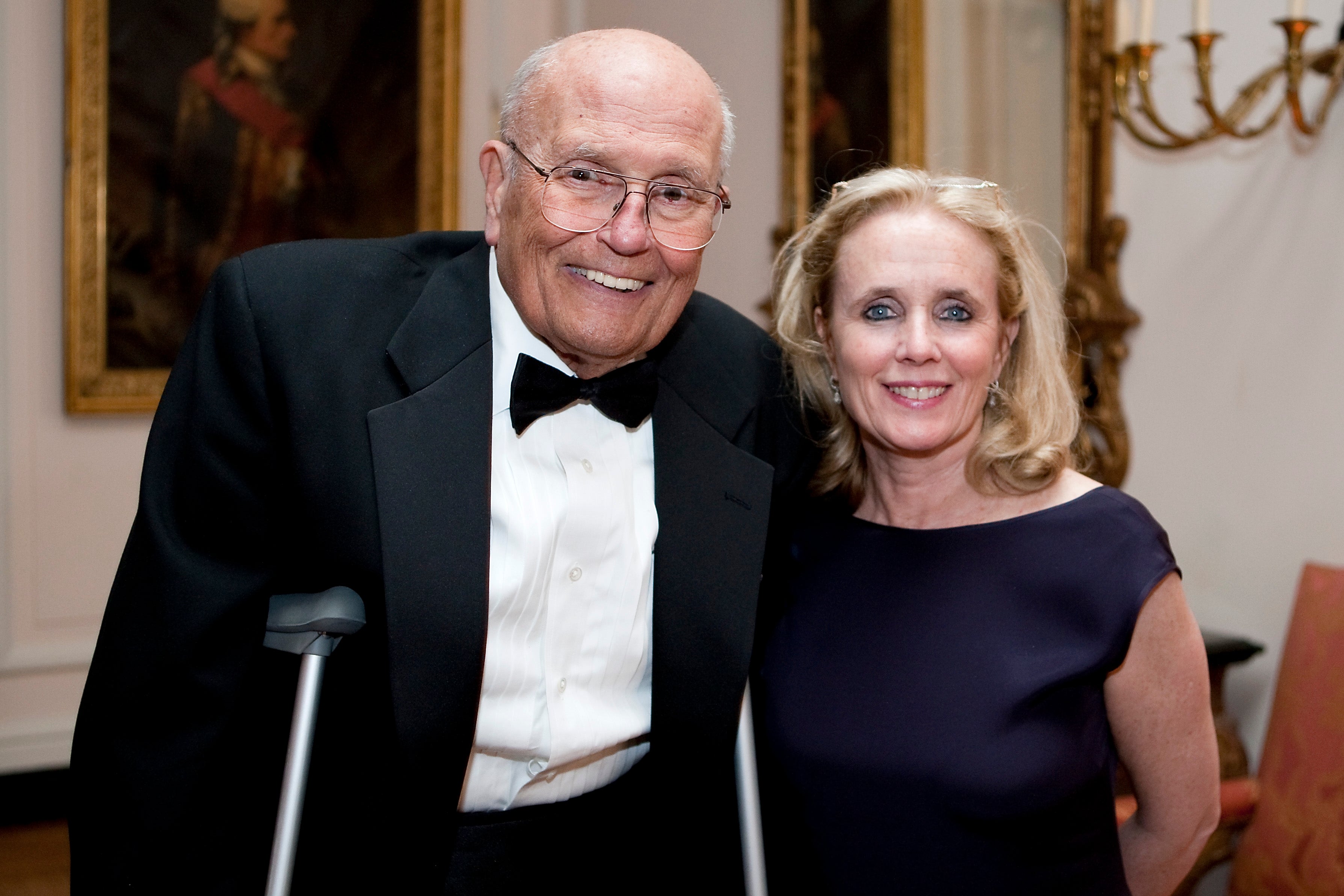Two people in formal attire, one in a tuxedo using crutches, in an elegant room with artwork and chandeliers