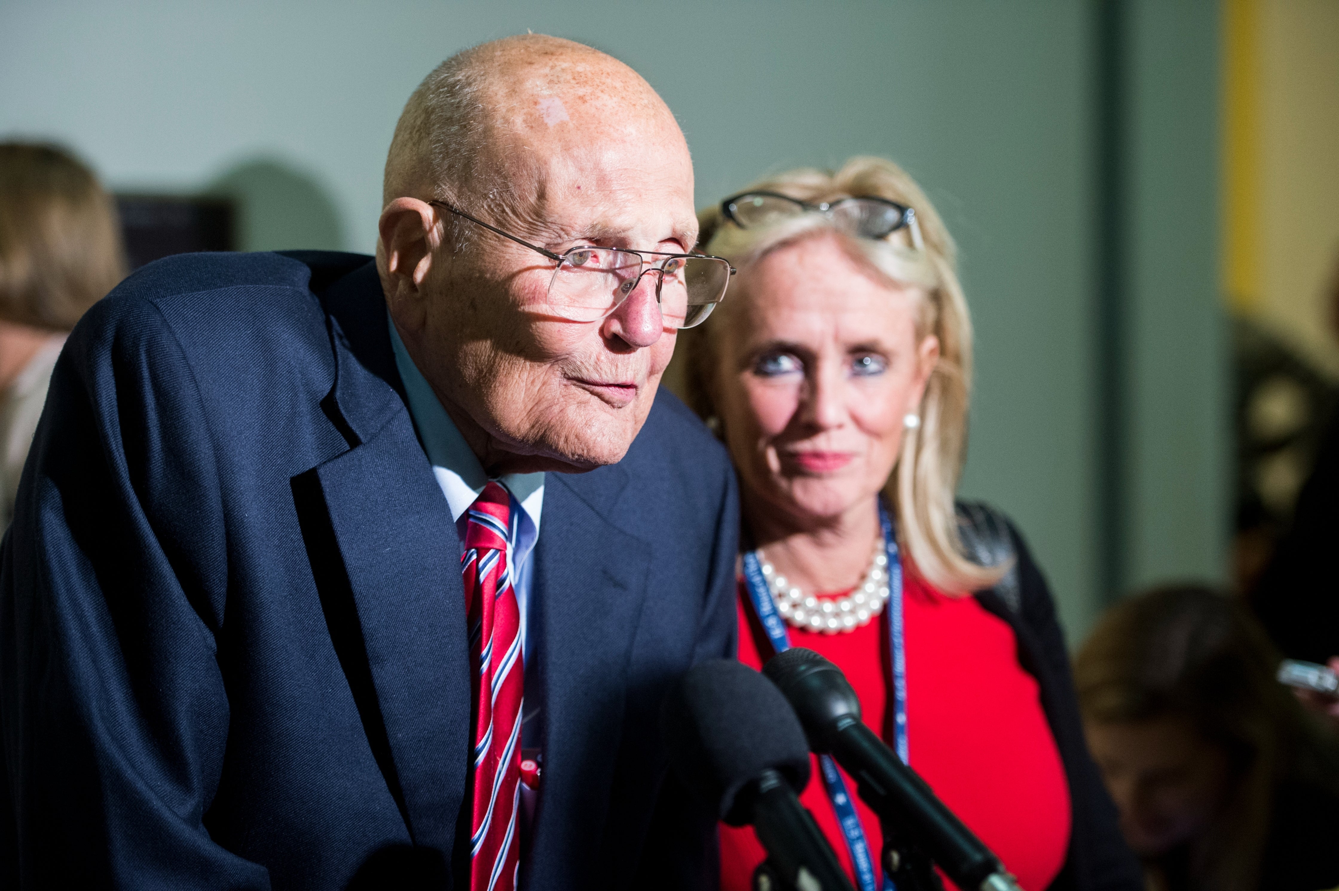 Elderly man and a woman stand together at a microphone during a public event. The man wears a suit and tie, the woman wears glasses and a necklace