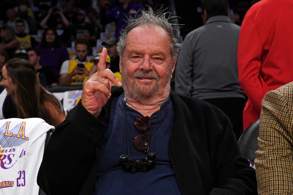 Jack Nicholson seated courtside at a basketball game, wearing casual attire and holding his index finger up