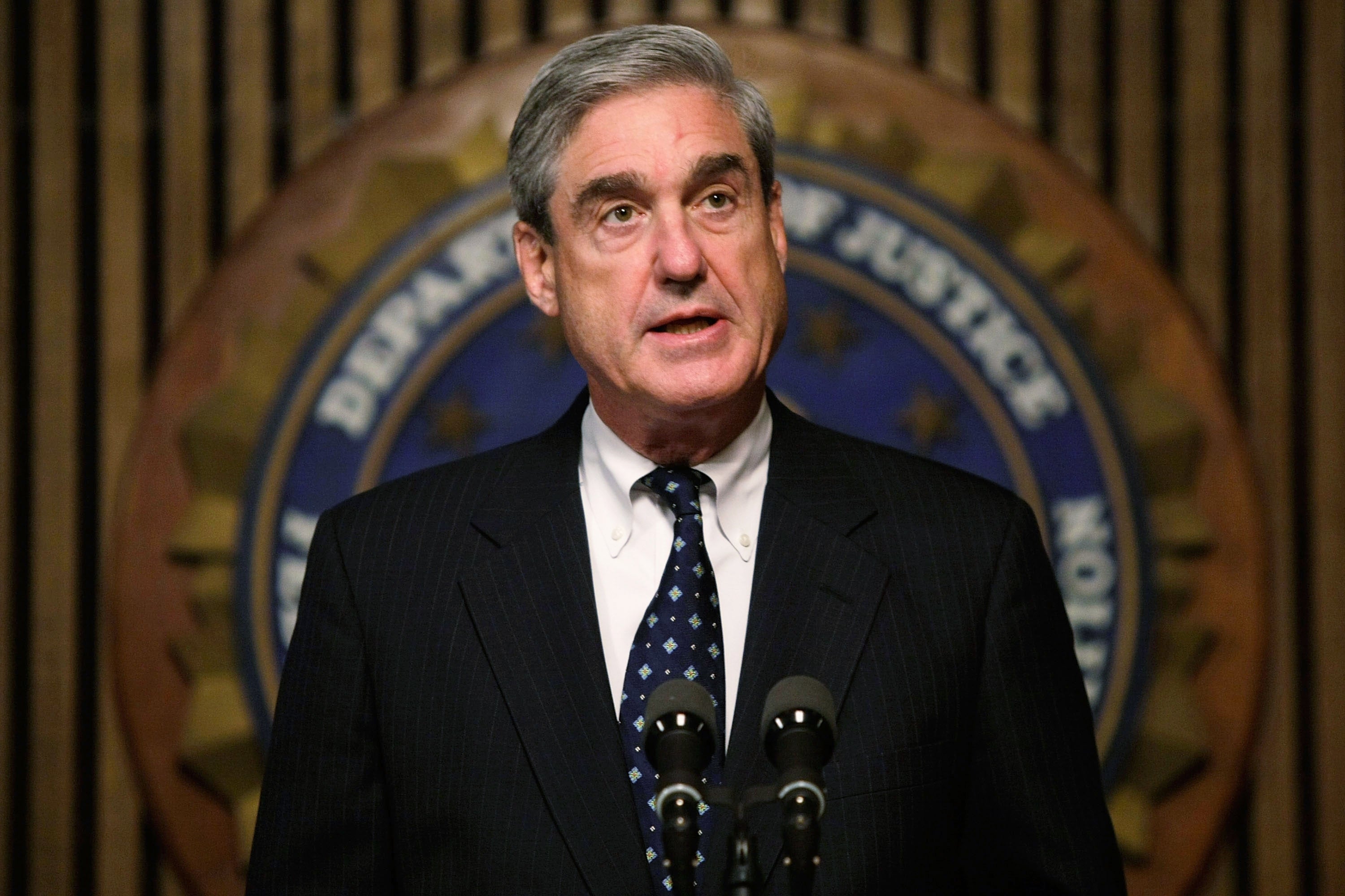 A person in formal attire speaks at a podium, standing in front of a Department of Justice emblem