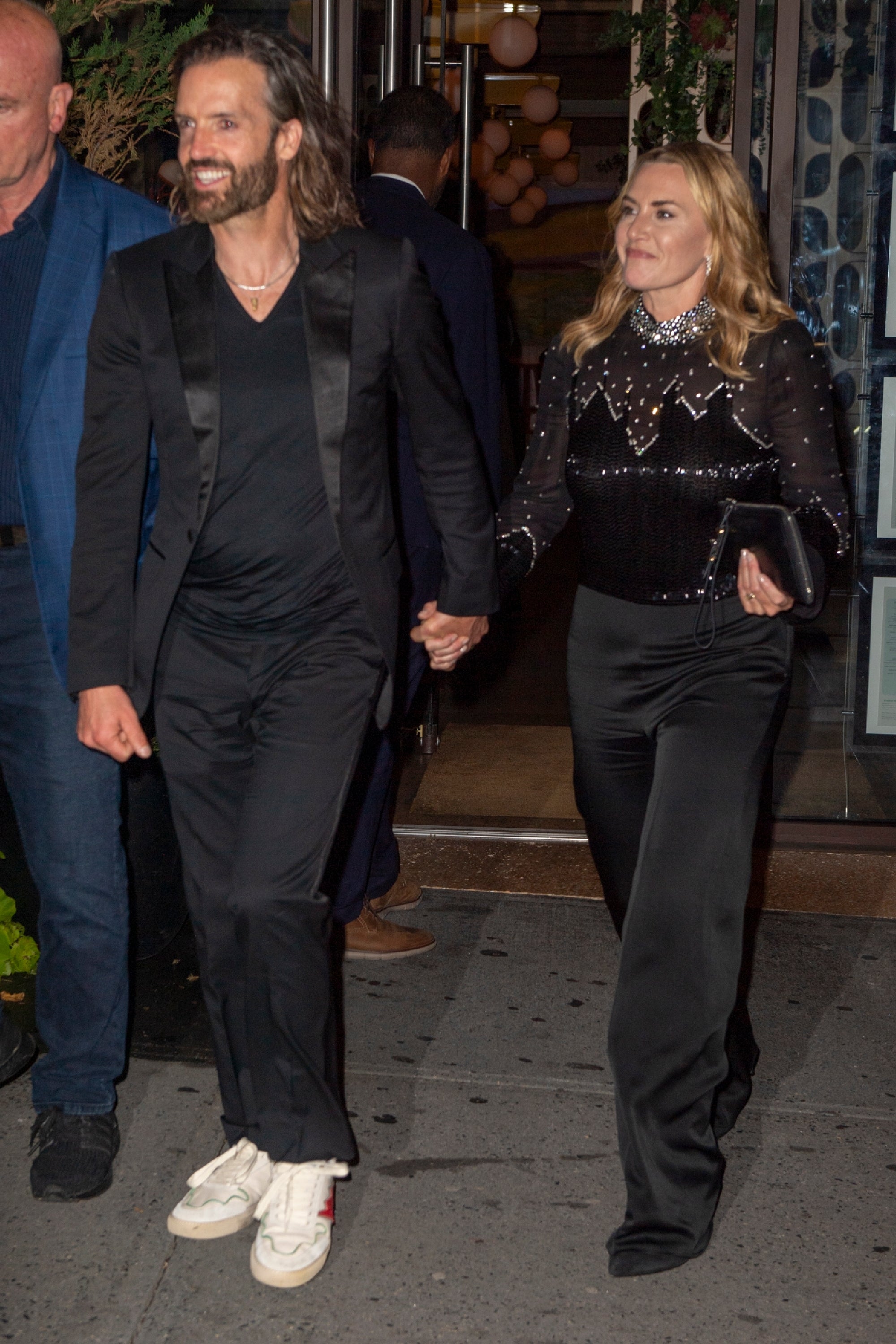 A man in casual attire and a woman in a sparkling top and sleek pants hold hands, walking outside, possibly attending a celebrity event