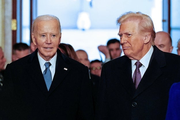 Two public figures in formal attire walk together at an event, engaging with attendees in the background
