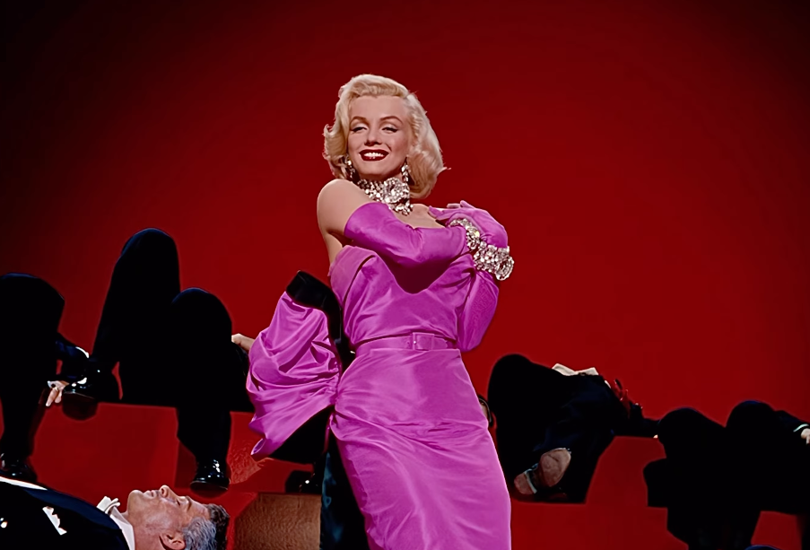 Person in elegant, vintage style outfit with gloves, standing on stage with a confident pose, surrounded by people lying down