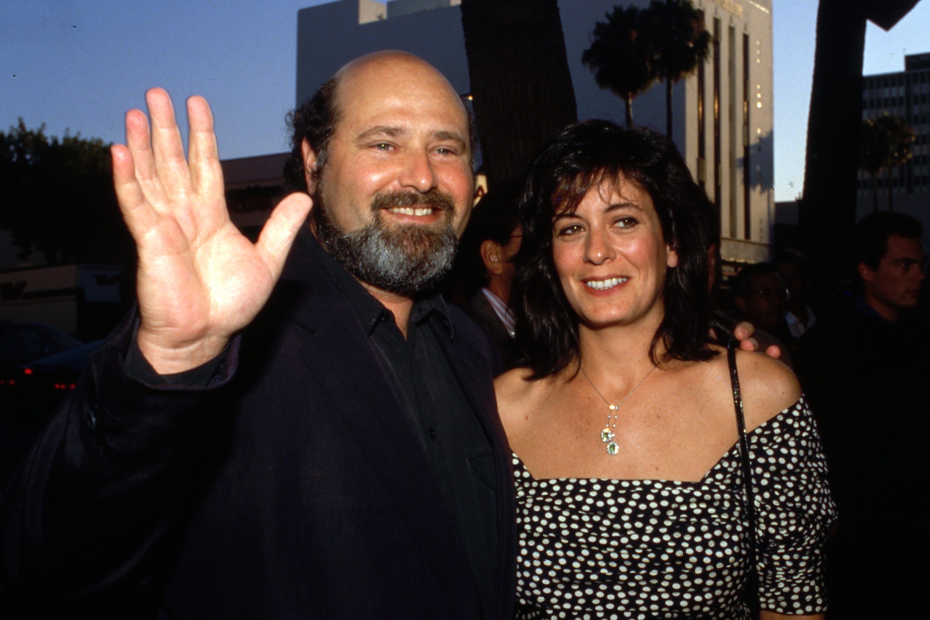 Two people smiling at an outdoor event. The man waves, wearing a dark suit; the woman wears an off-shoulder polka dot dress