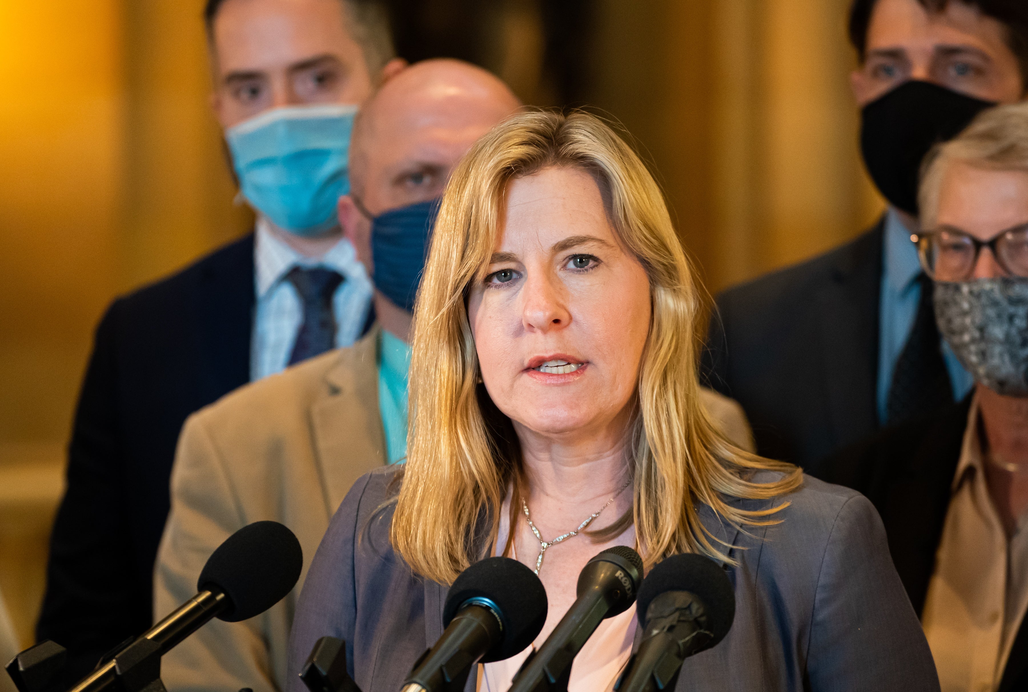 A woman speaks at a press conference, surrounded by people, some wearing masks