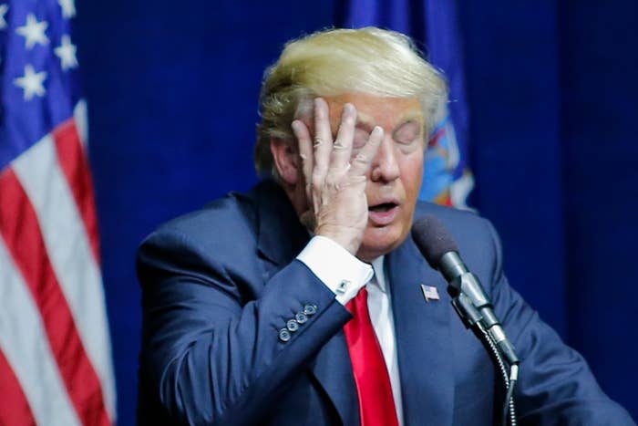 A person in a suit stands at a podium with a flag behind them, covering their face with one hand, appearing frustrated or contemplative