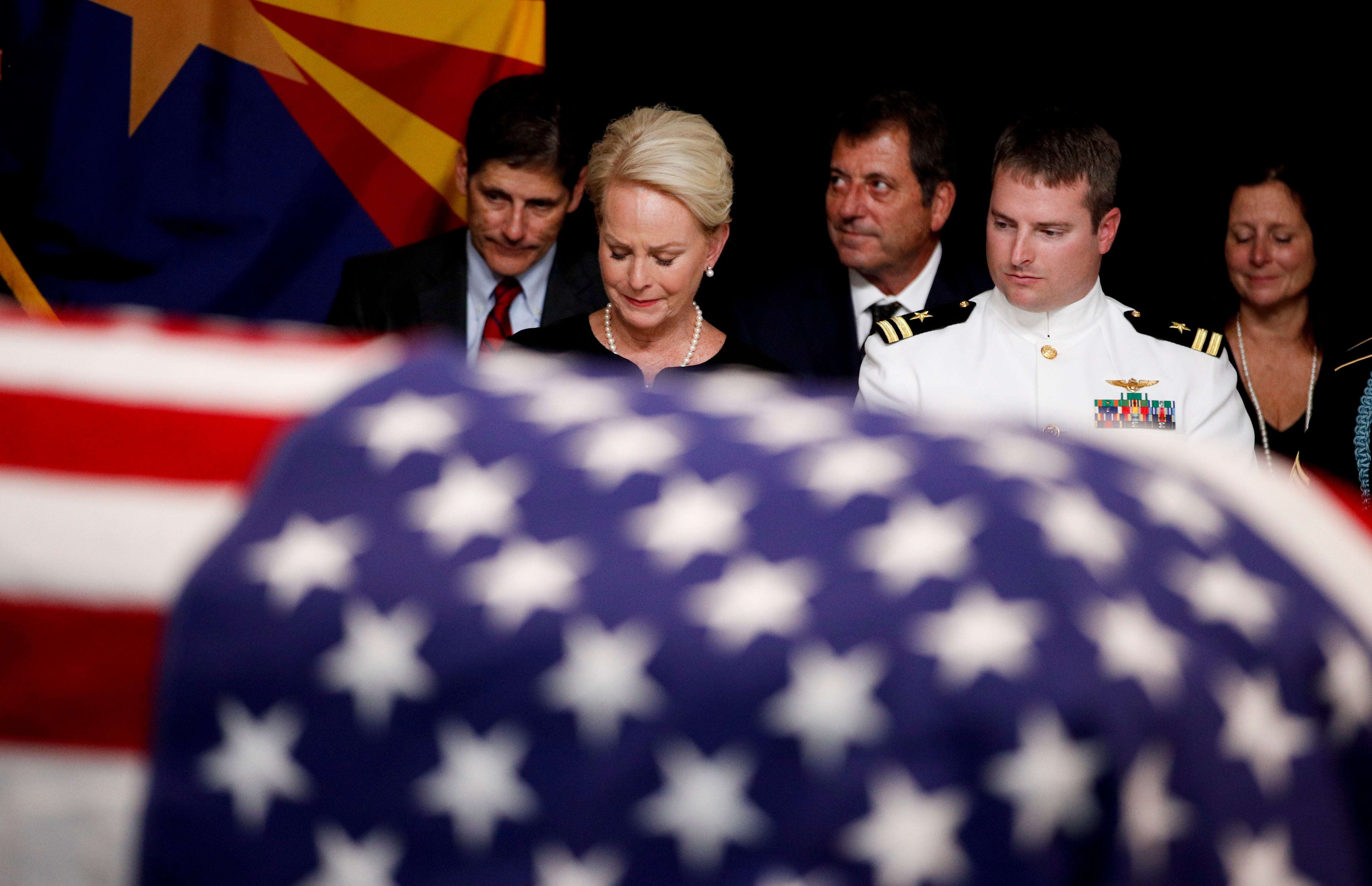 A group of people stand solemnly beside an American flag-draped casket in a ceremonial setting