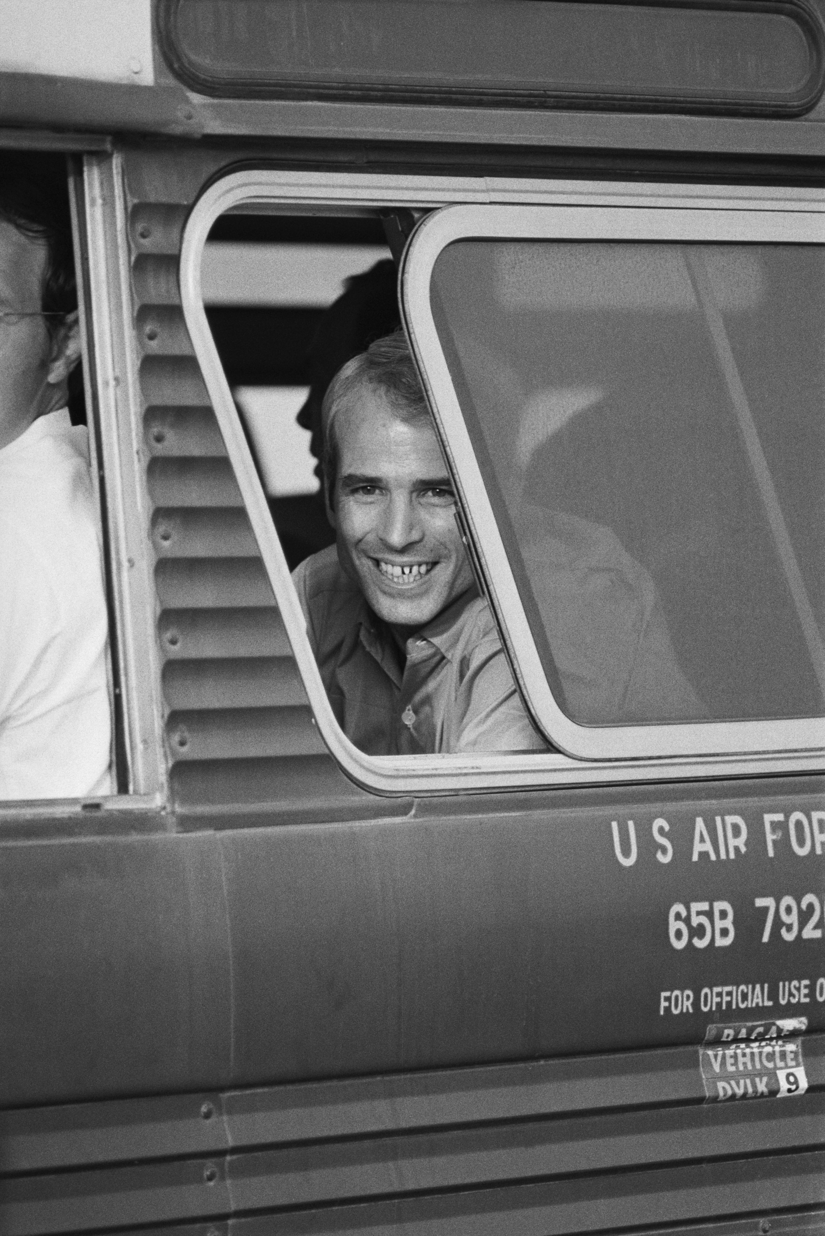A man smiles broadly while looking out a bus window labeled "U.S. Air Force," seated among others inside