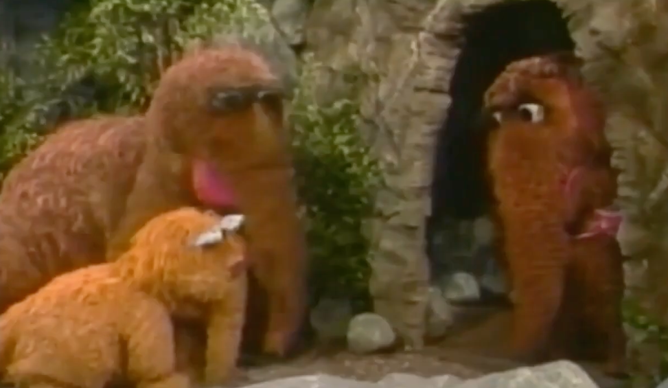 Three large, furry puppet characters interact in a cave-like setting, reminiscent of a vintage children's show scene