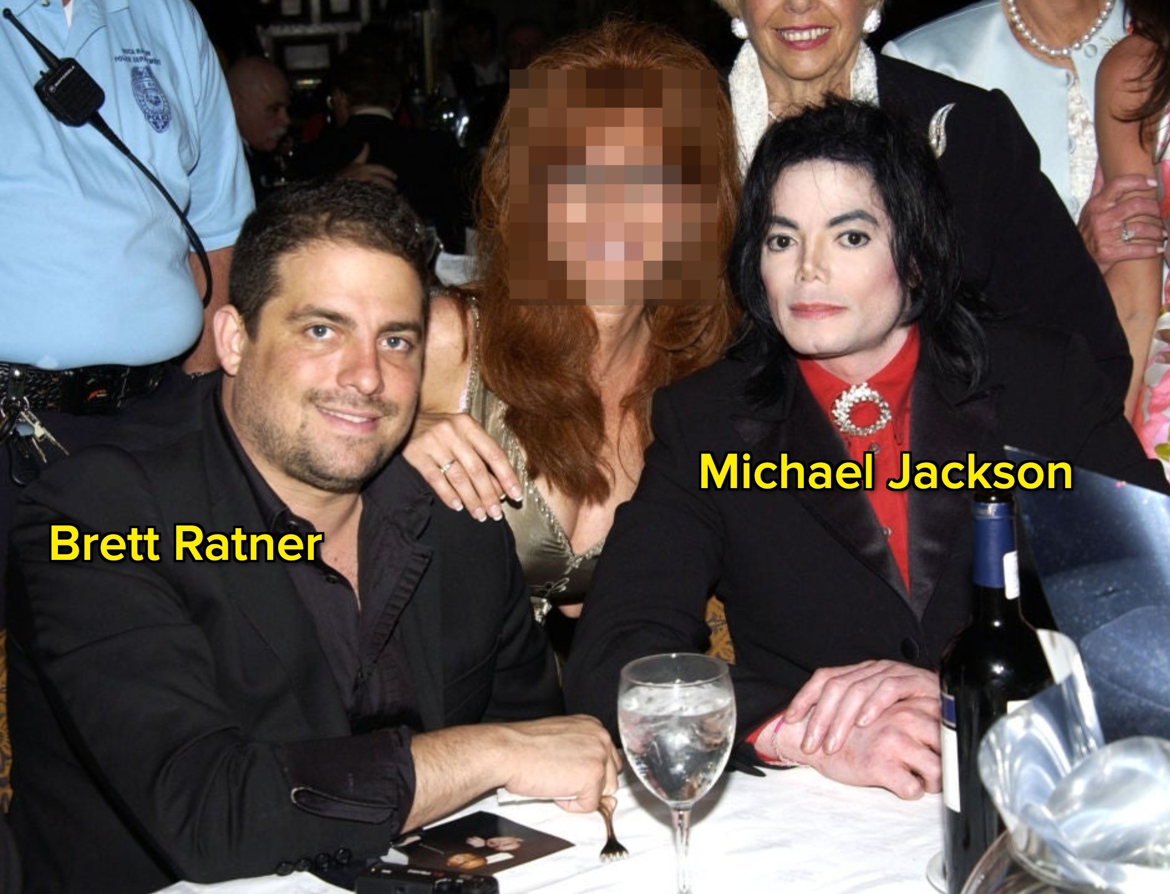 Group of people at an event, with one person in a red shirt and black blazer. They are gathered around a table with a bottle and glasses