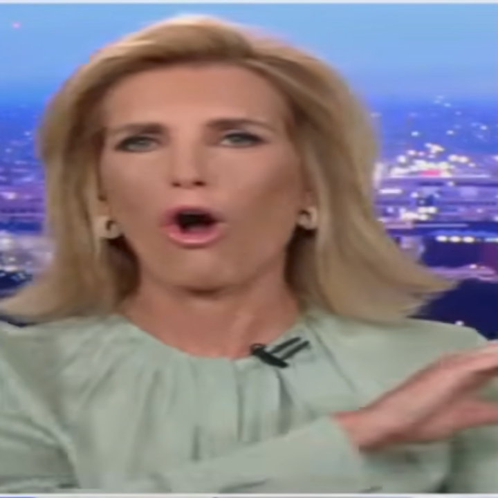 A person speaking on TV with a cityscape backdrop. They wear a light blouse and gesture while talking