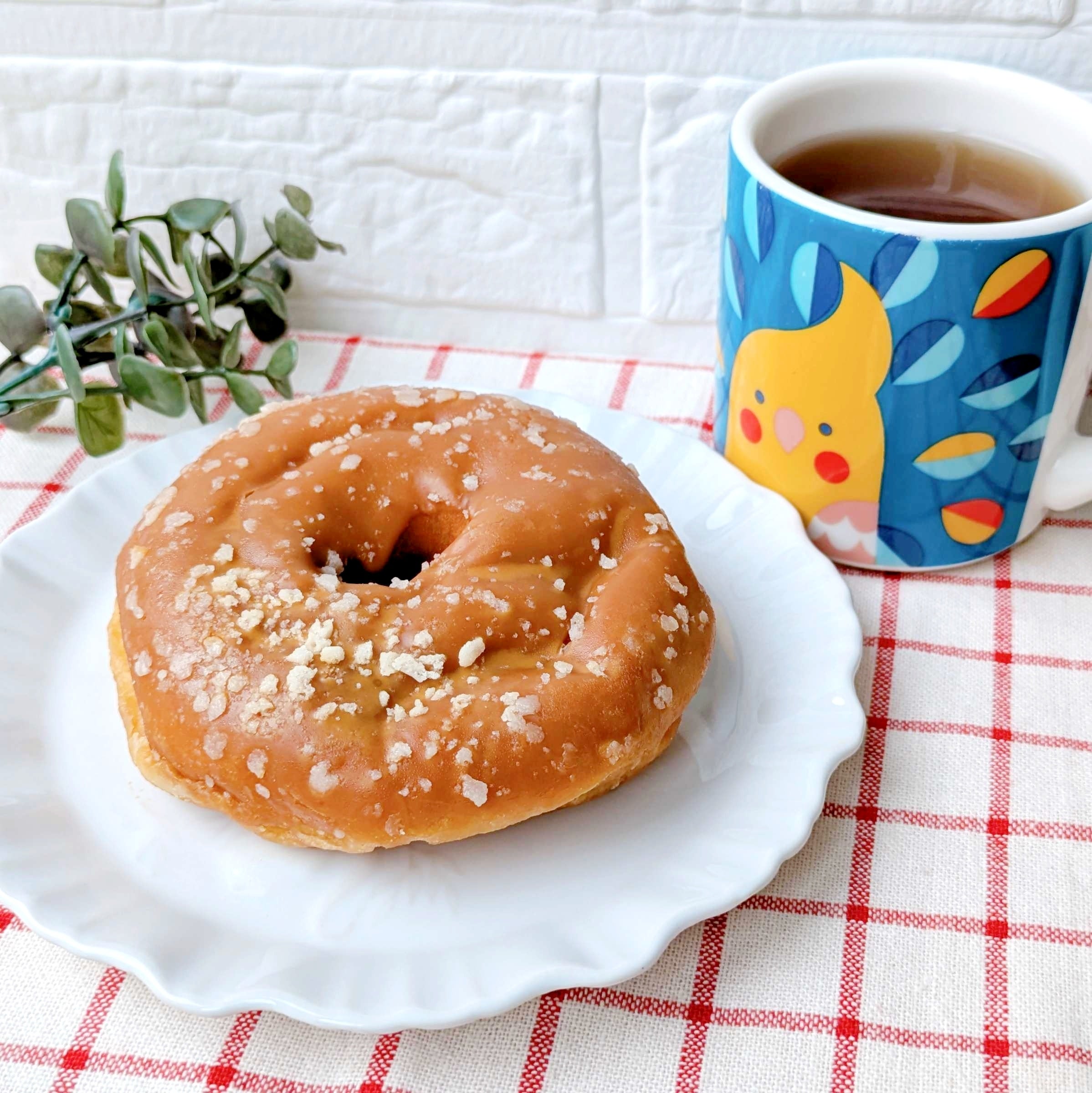ファミリーマート「も～っちり食感きなこドーナツ」ドーナツと植物の置物、デザインがプリントされたマグカップに入った飲み物がチェック柄のテーブルクロスに置かれている。