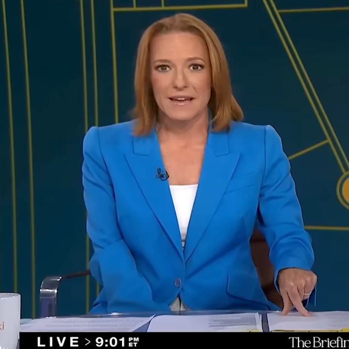 A person in a blue blazer sits at a news desk, appearing to deliver a broadcast