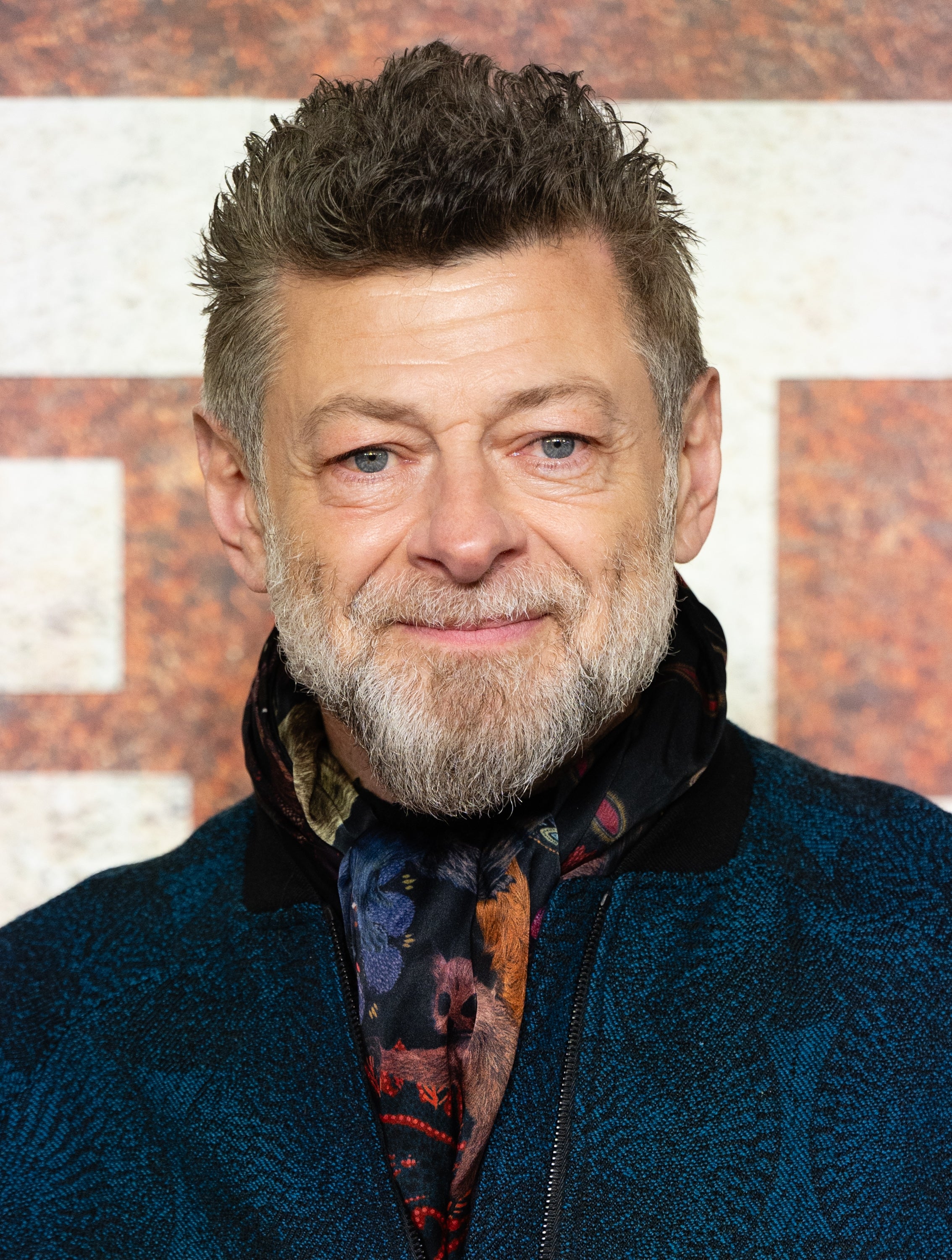 A person with short, styled hair and a beard, wearing a patterned scarf and textured jacket at a public appearance