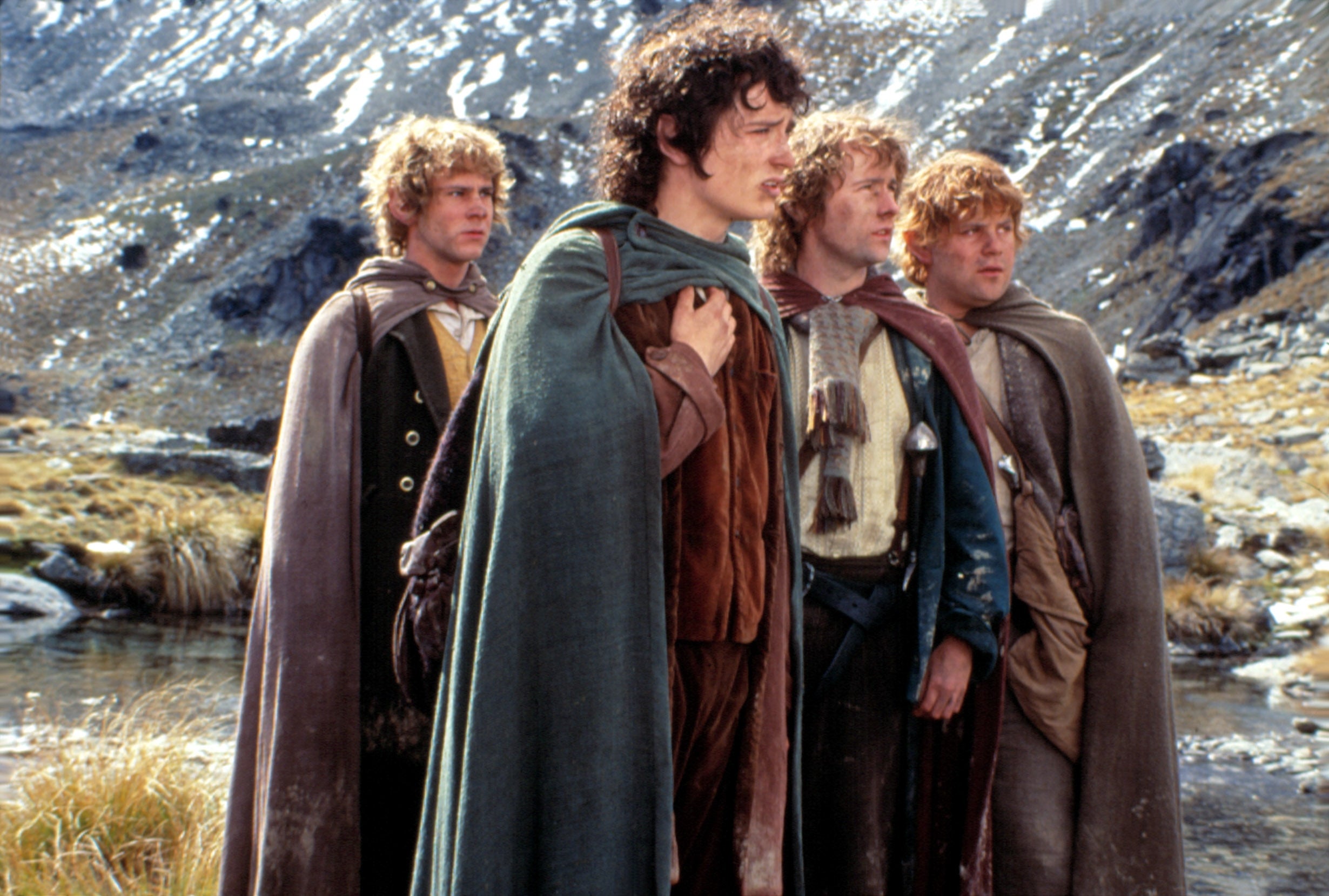 Four actors in medieval-style costumes stand outdoors beside a creek, appearing focused and contemplative