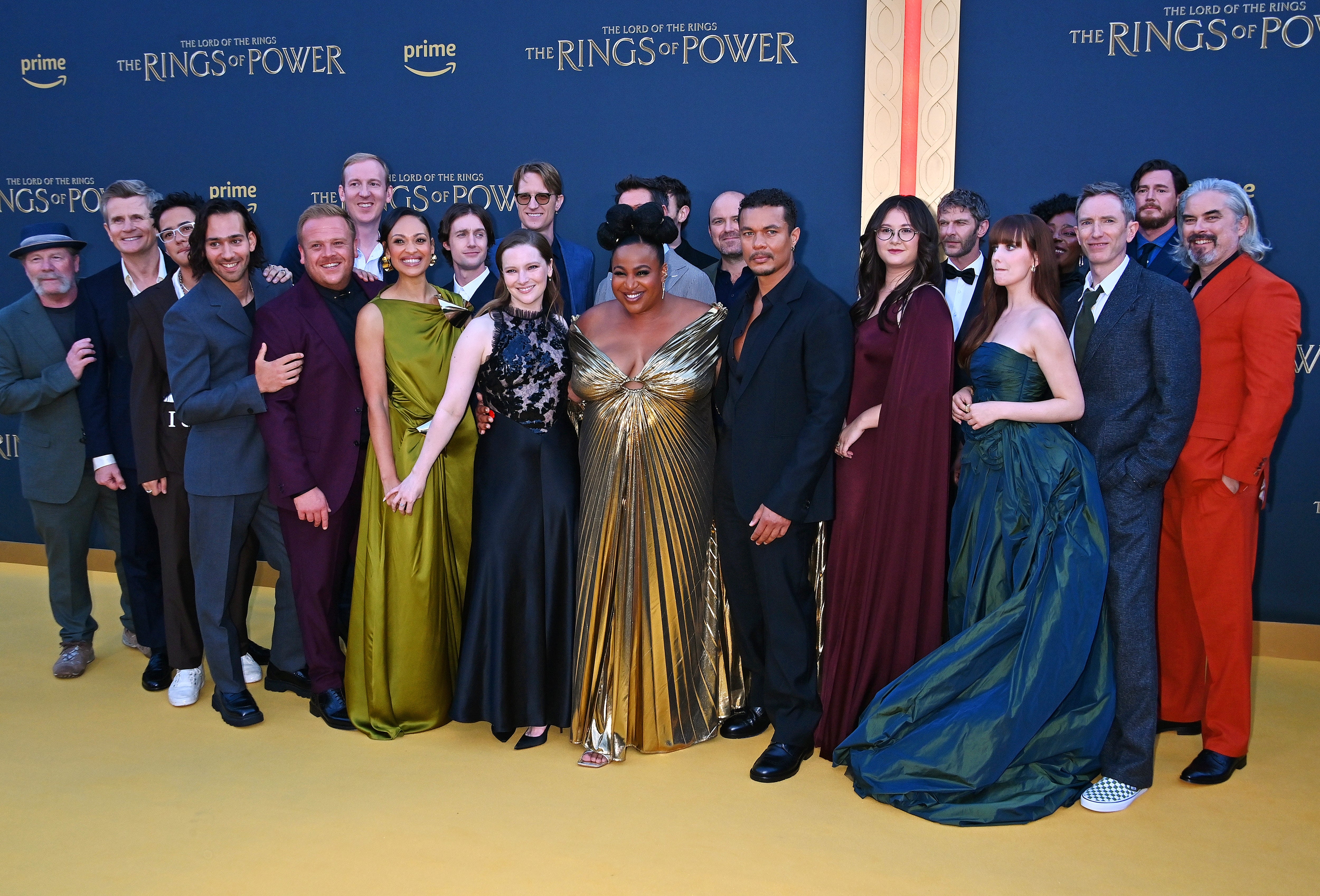 A group of actors pose on a red carpet for "The Rings of Power." They wear a mix of formal suits and elegant gowns